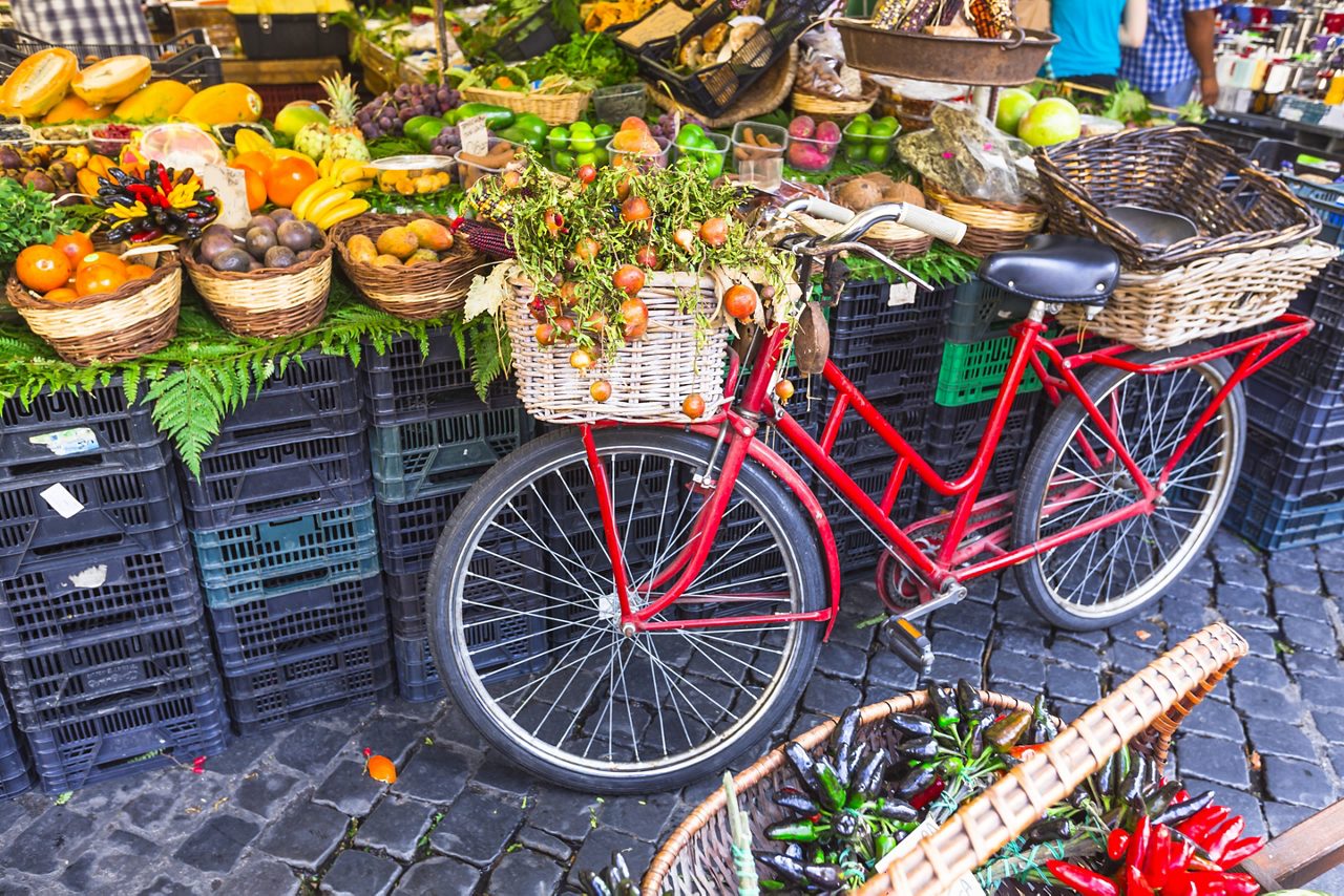 A red bicycle with baskets rests against milk crates holding fruits and vegetables.