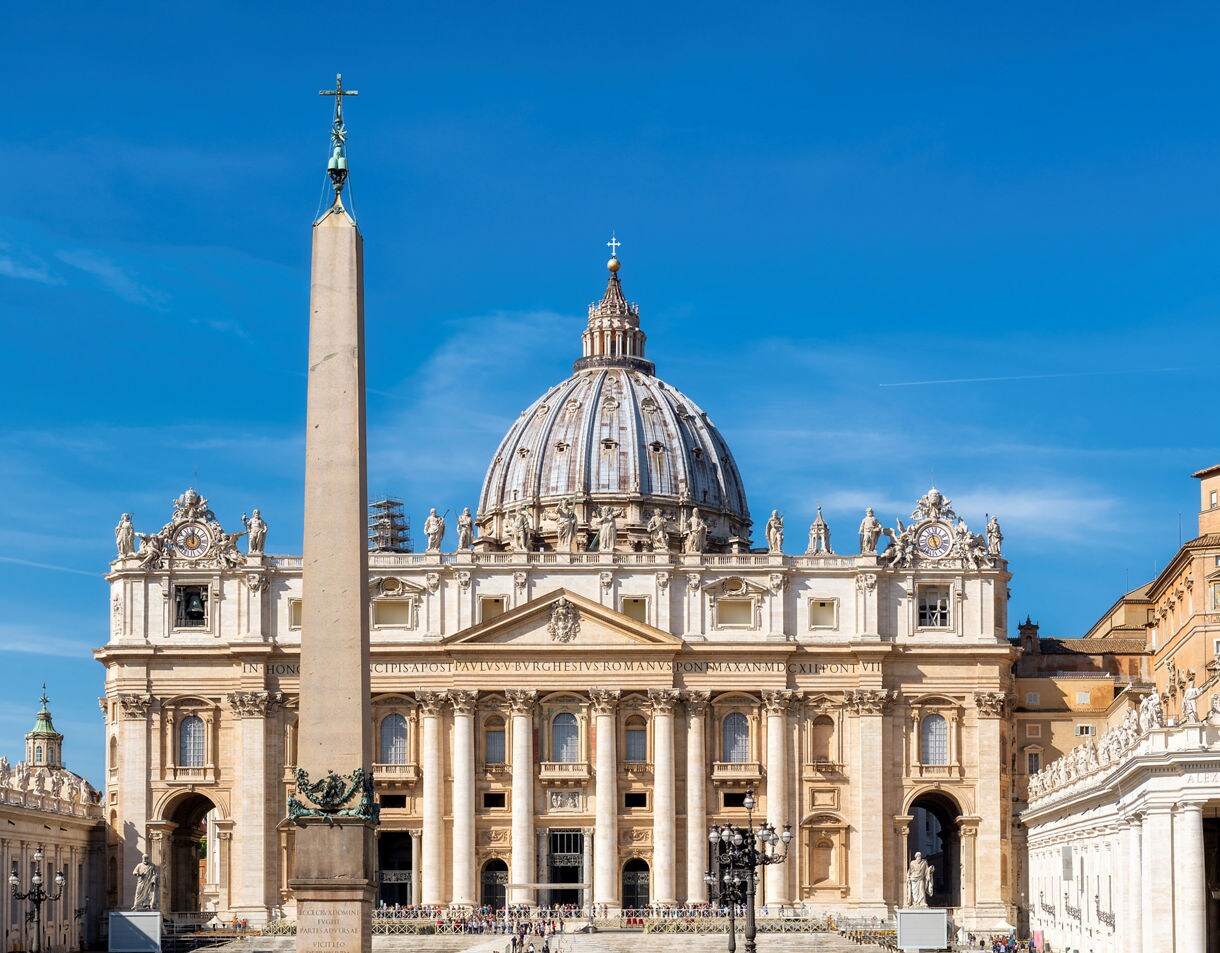 Front view of St. Peter’s Basilica with its dome and central obelisk under a bright blue sky.