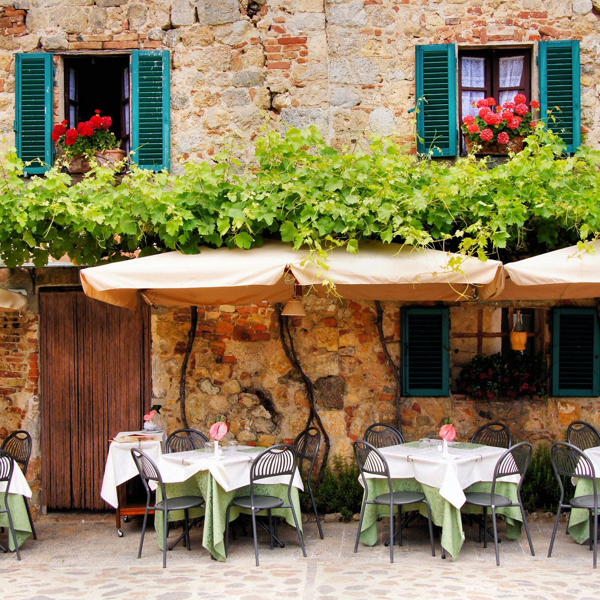 Outdoor Italian trattoria with stone walls, green shutters, vine-covered awnings, and tables set for dining.