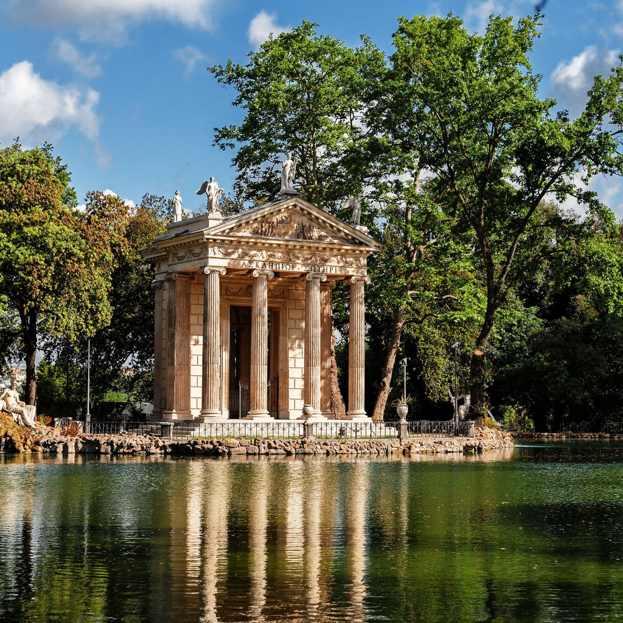 Neoclassical temple with columns reflected in a calm lake, surrounded by green trees in Villa Borghese.