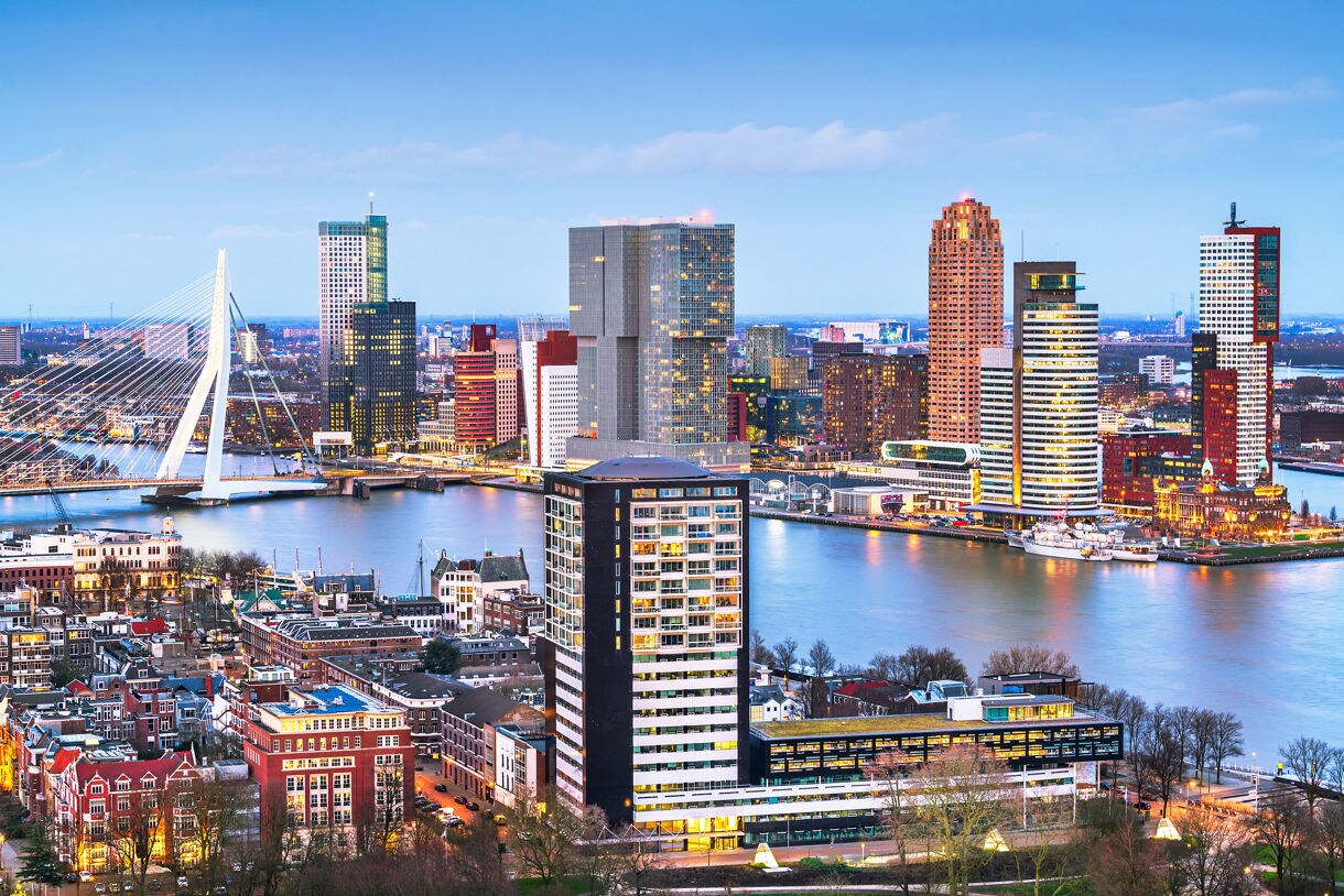 Evening view of Rotterdam, Netherlands, featuring the illuminated skyline, Erasmus Bridge and high-rise buildings along the waterfront.