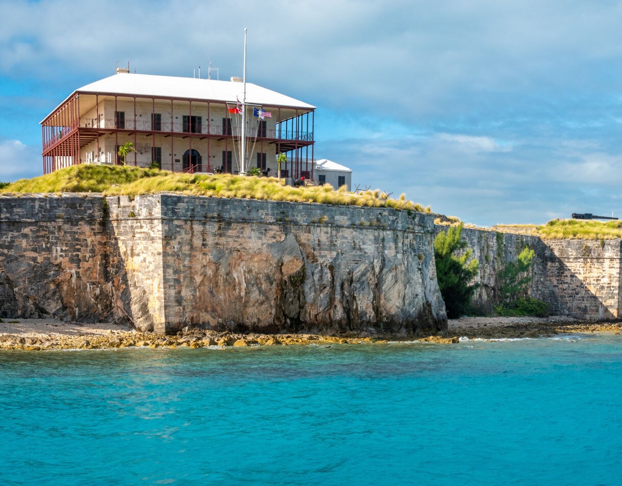 Historic stone fort with grassy ramparts and a colonial building above, overlooking bright blue waters at the Royal Naval Dockyard in Bermuda.