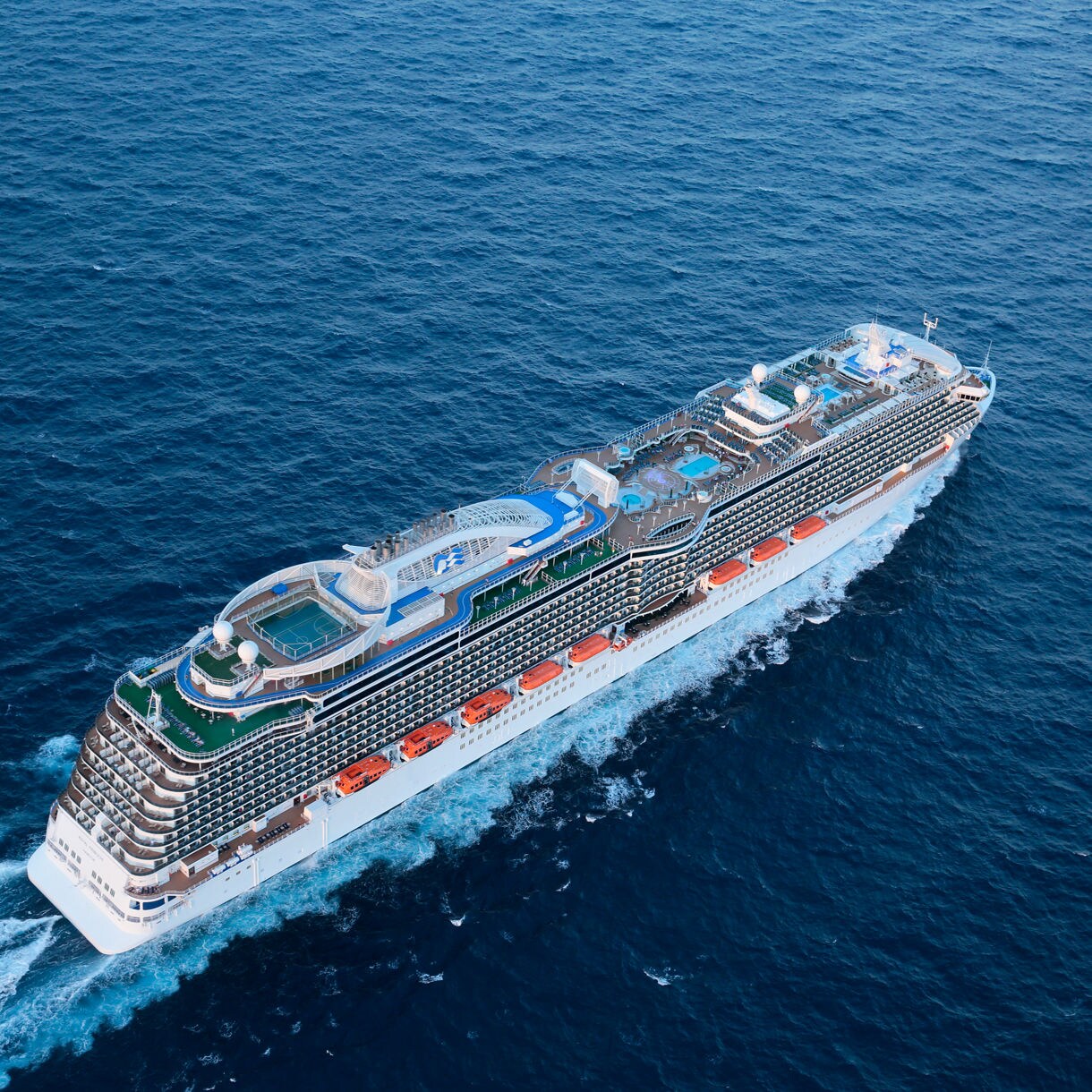 The image shows a brightly lit cruise ship captured from above as it moves through dark waters at night. Vivid blue and white waves trail behind the vessel, creating a striking visual contrast. The ship's deck is illuminated with warm lights, and a swimming pool is visible at the center. The overall scene conveys a sense of luxury, travel, and nighttime adventure.