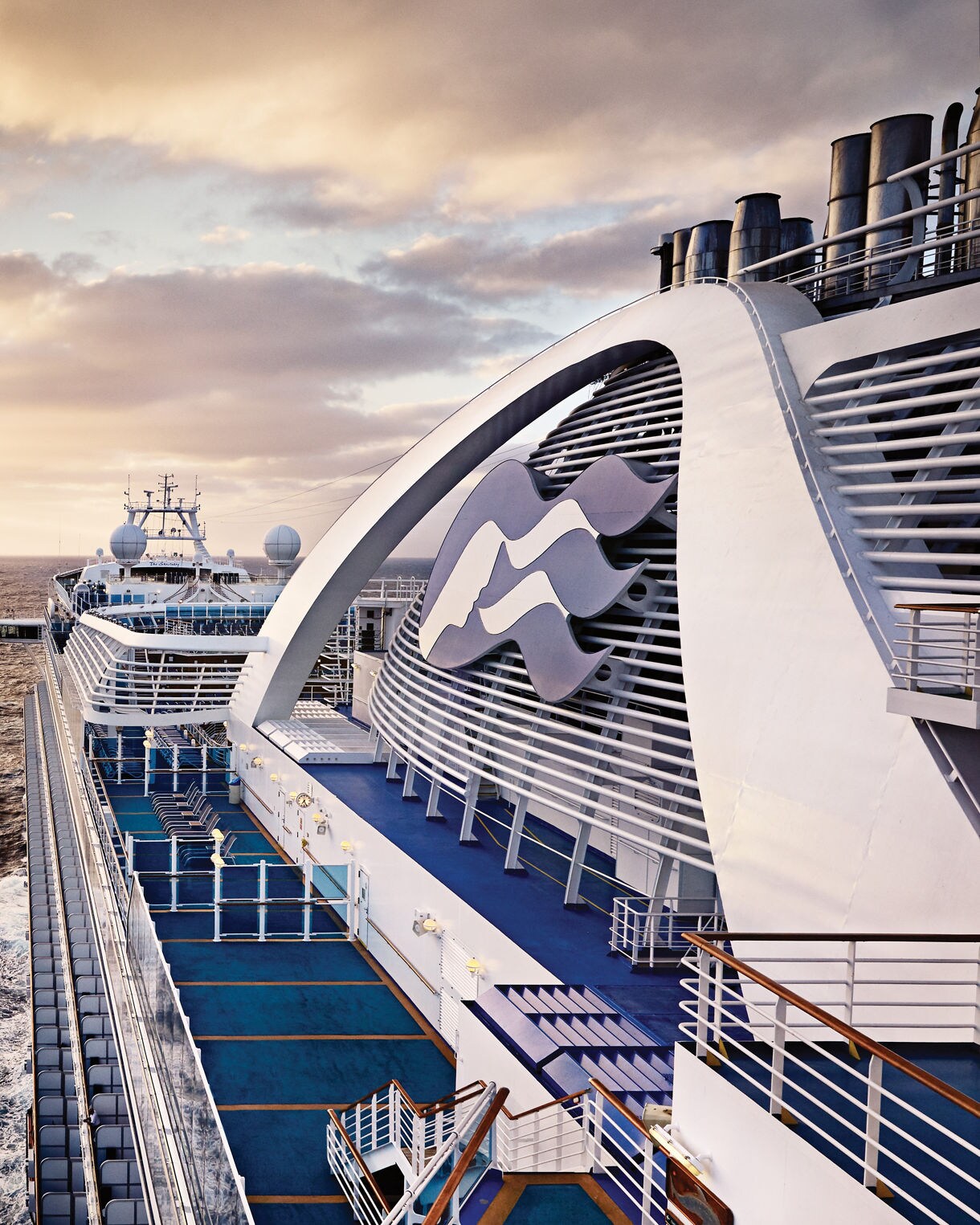 A large modern cruise ship travels across a vast open ocean at sunset. The camera angle looks along the upper decks, showing railings, pools, and outdoor areas stretching toward the stern. Soft golden light from the setting sun reflects on the water and the ship’s surfaces, creating a warm, tranquil atmosphere. The sky is partly cloudy, adding depth and drama to the seascape.