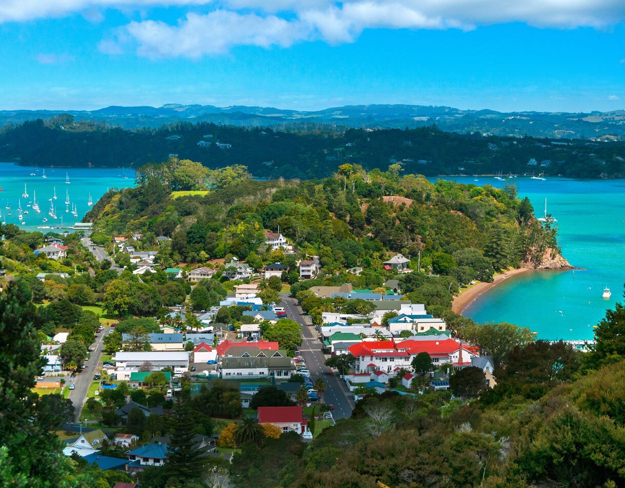 Aerial view of Russell, New Zealand, showing colorful seaside houses, lush hills and turquoise waters dotted with sailboats.