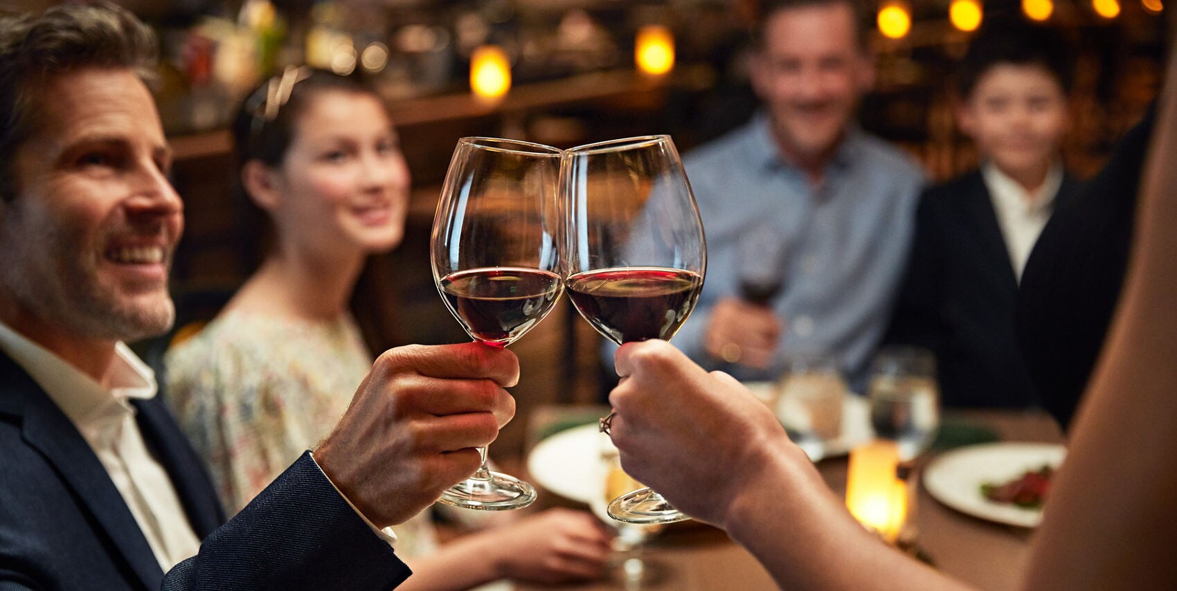 Two people clinking red wine glasses at a candlelit dinner table with smiling guests in the background.
