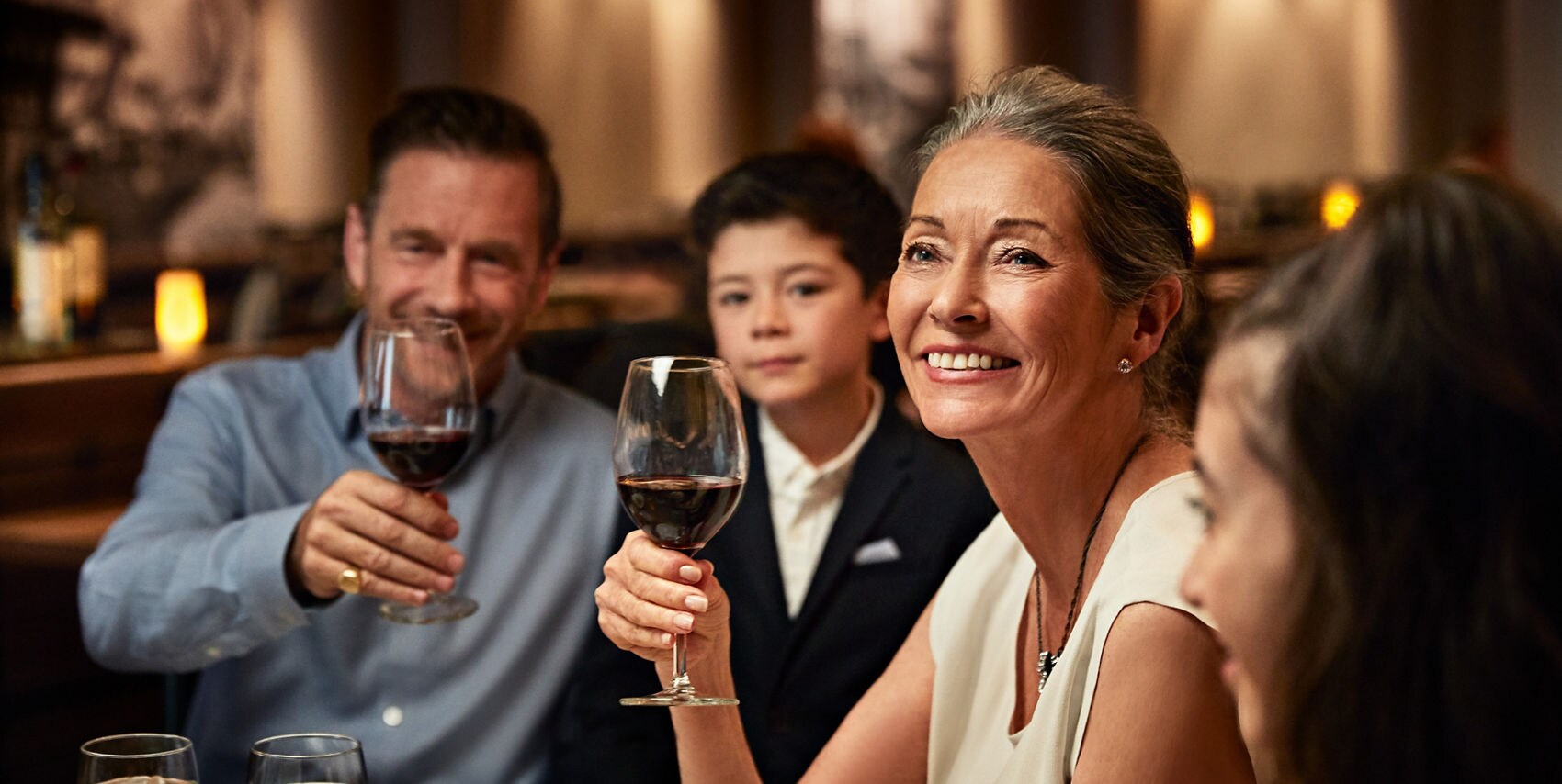 A group of adults enjoying a social gathering in a warmly lit restaurant setting. They are holding glasses of red wine, suggesting a celebratory or casual dining atmosphere. The table is set with water glasses and plates, enhancing the fine dining ambiance.