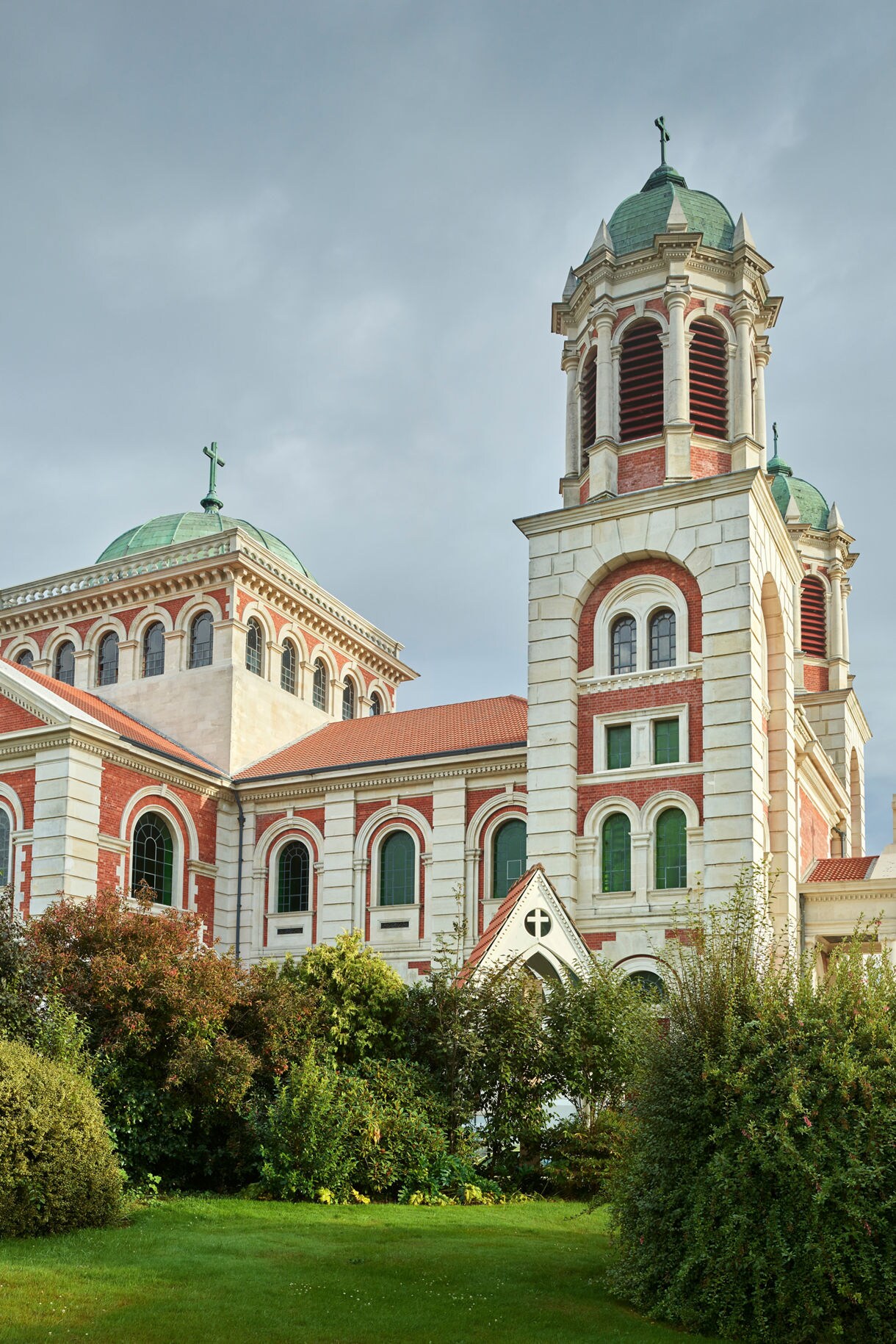 Large basilica with red-and-white brickwork, green domes and arched windows framed by lush garden shrubs under a cloudy sky.