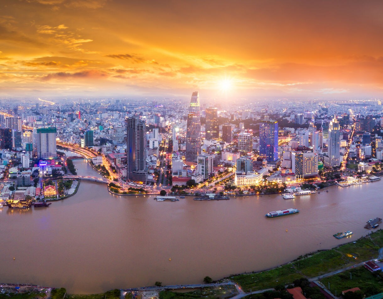 Aerial view of Ho Chi Minh City at sunset, showing the Saigon River winding through illuminated skyscrapers beneath a glowing orange sky.
