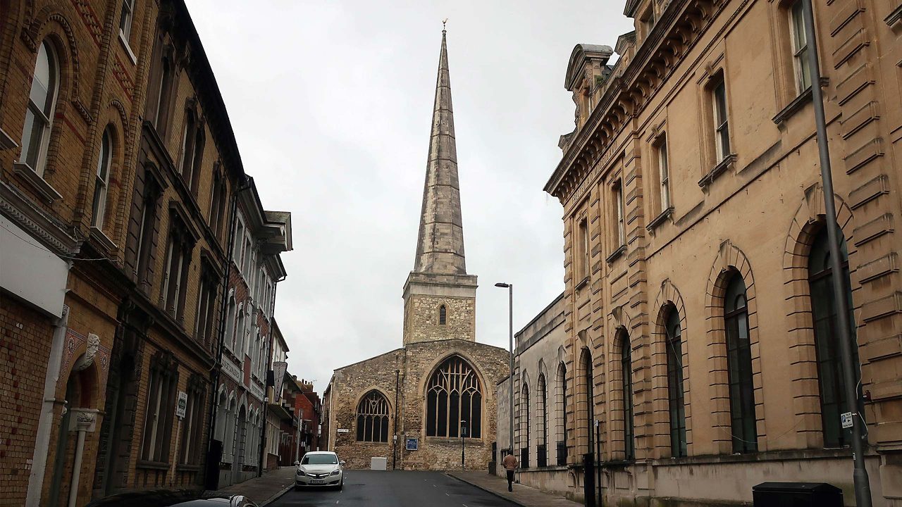 St. Michael's Church in Southampton, England, showcasing its tall stone tower and pointed spire. The church's historic architecture stands out with its arched windows and weathered stonework, set against a bright sky. The surrounding area appears quiet, highlighting the church as a prominent feature in the neighborhood.