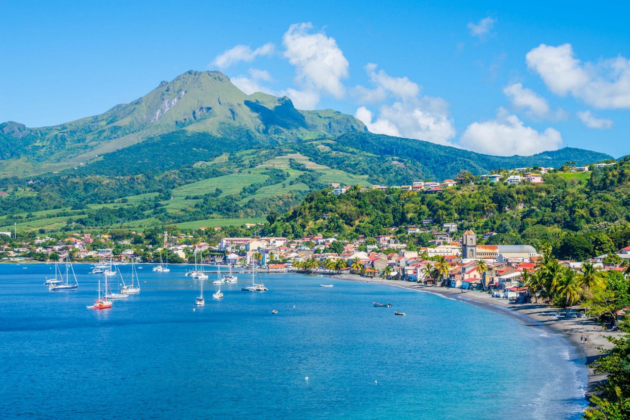 Scenic harbor in Martinique with white sailboats anchored in blue bay, colorful waterfront buildings, and dramatic mountain backdrop.