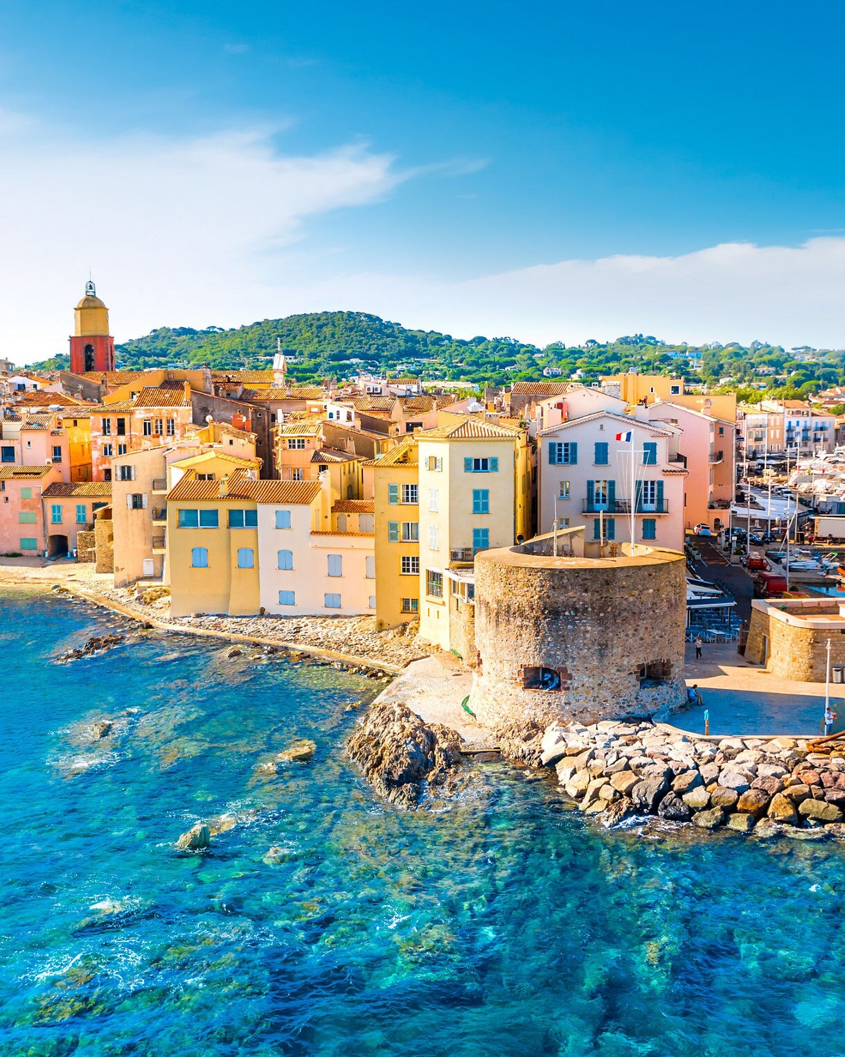 A coastal view of Saint-Tropez with pastel buildings lining the waterfront, a stone tower by the sea and a marina filled with boats, surrounded by bright blue water and green hills.