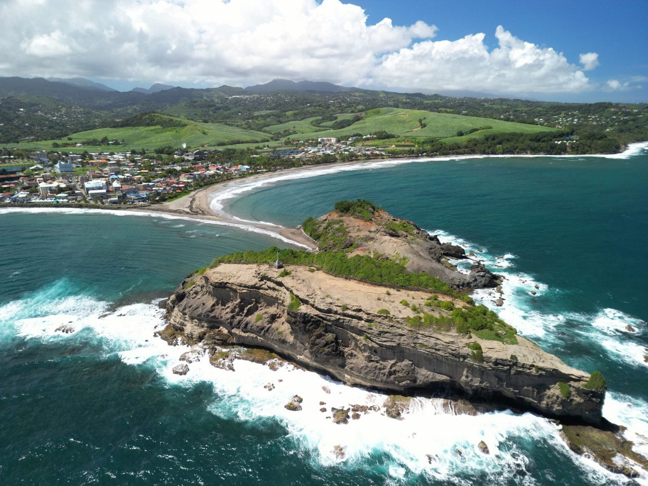 Aerial view of rocky peninsula jutting into turquoise Caribbean waters with coastal town and green mountains in background in Martinique.