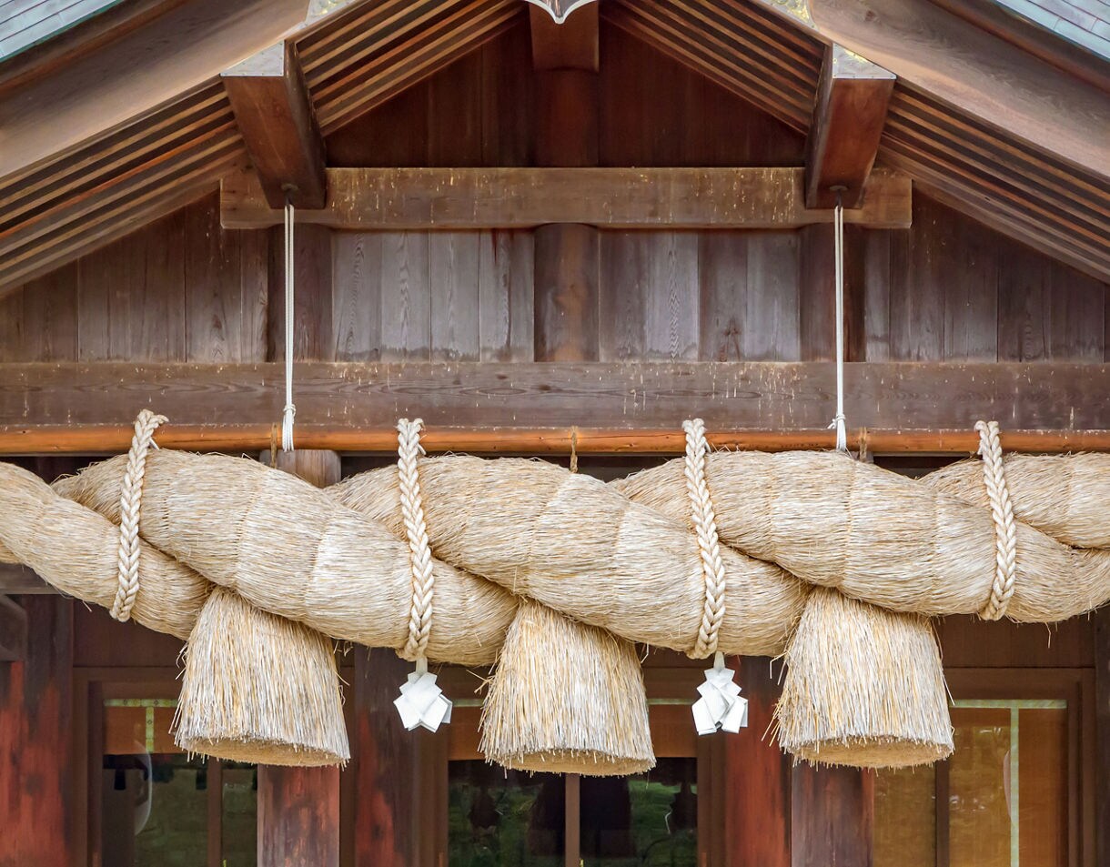 Close-up of a giant straw rope (shimenawa) hanging under the eaves of Izumo Grand Shrine, flanked by traditional lanterns.