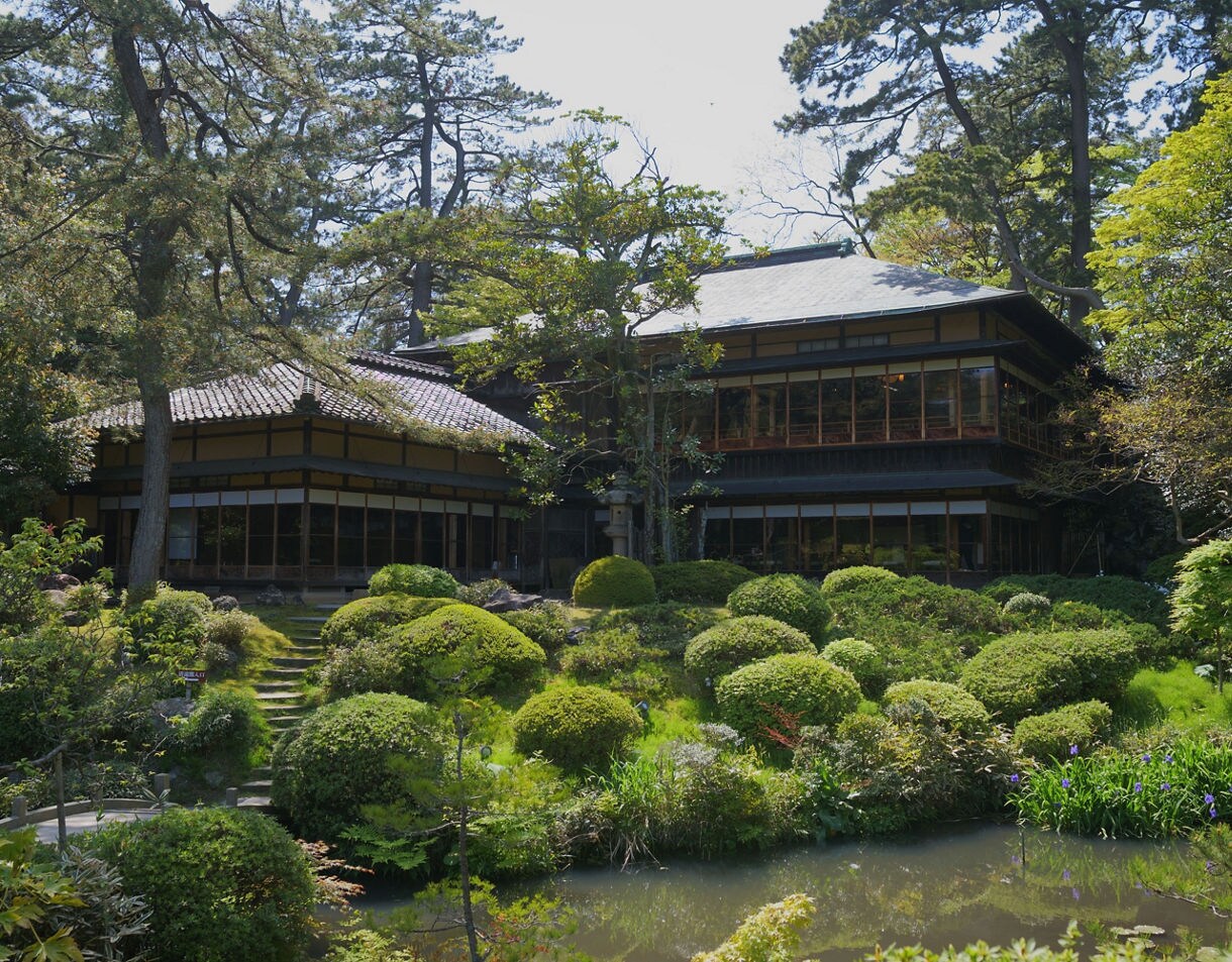 Traditional Japanese building with tiled roofs surrounded by manicured garden shrubs and a small pond.