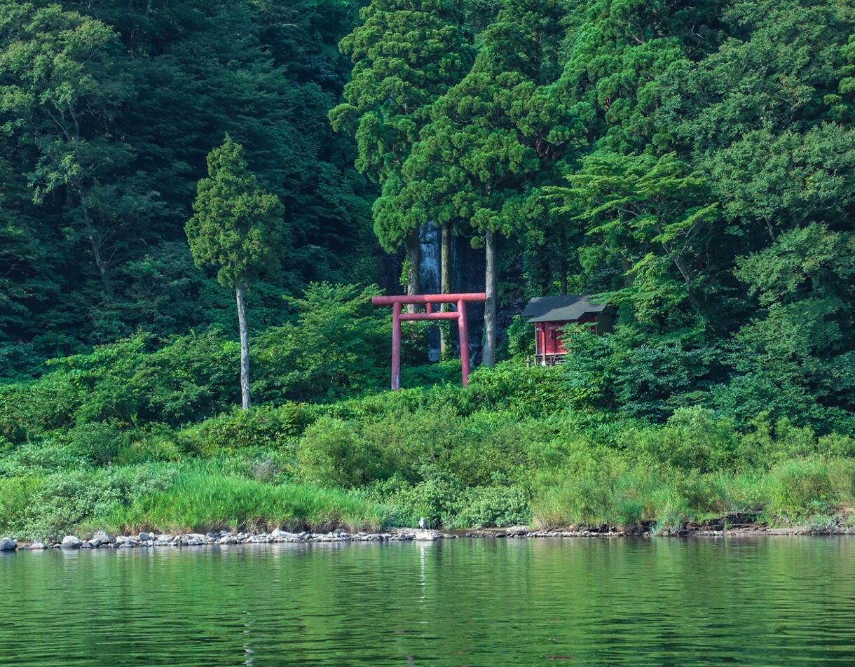 Red torii gate and small shrine nestled in dense greenery along the edge of the Mogami River.