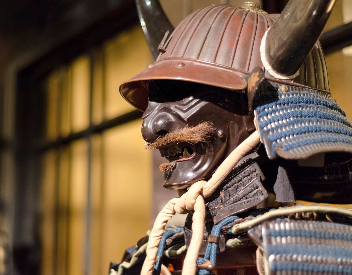 Close-up of traditional Japanese samurai armor featuring a horned helmet, dark mask and blue lacing.