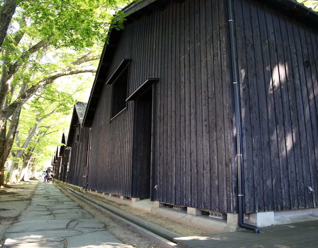 Row of dark wooden storehouses beside a stone walkway shaded by leafy green trees in Sakata, Japan