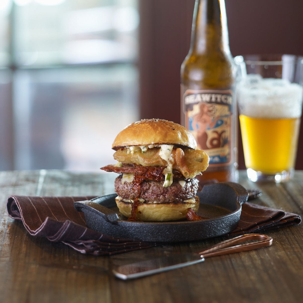 A gourmet burger on a brioche bun with melted cheese and toppings, served on a dark plate alongside a craft beer bottle and glass.