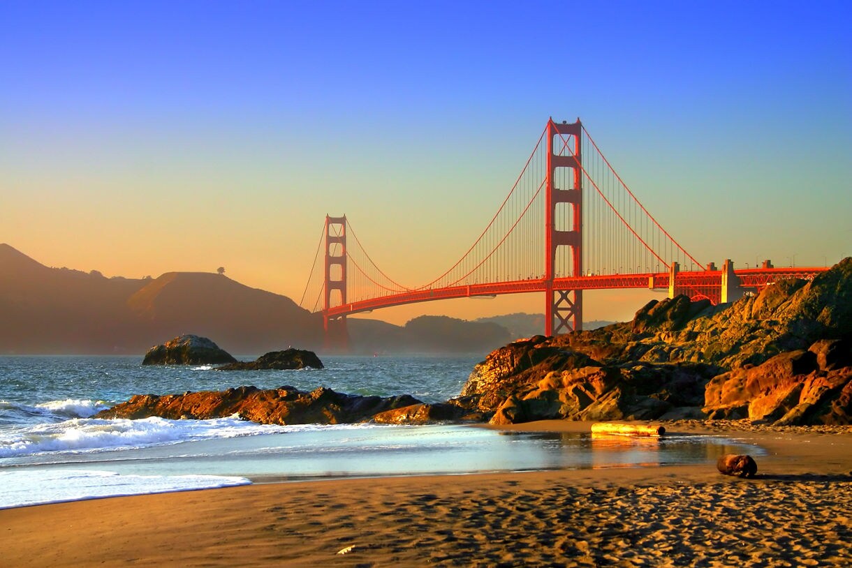 The iconic red-orange Golden Gate Bridge spans the San Francisco Bay, its massive suspension towers and sweeping cables standing against the backdrop of California's coast.