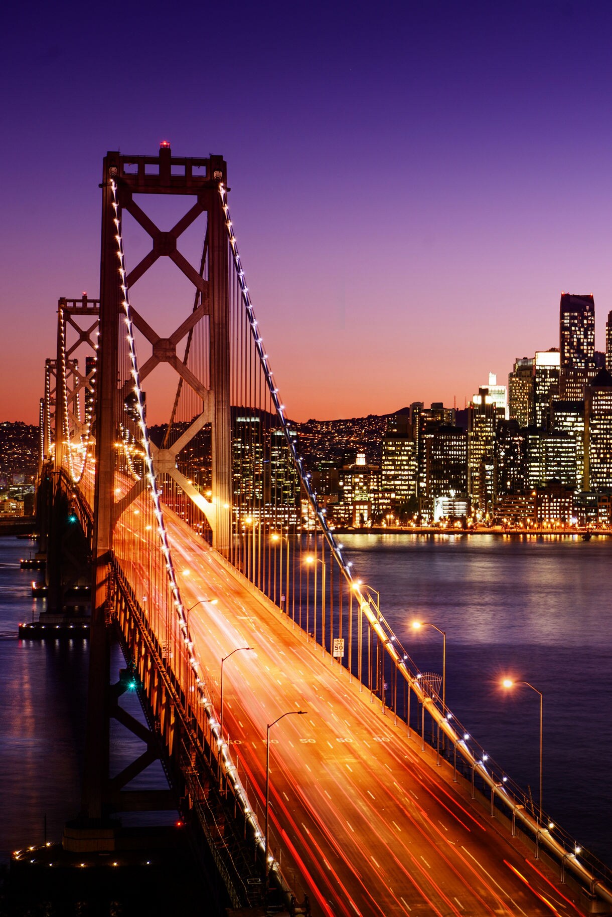 A nighttime view of the Bay Bridge in San Francisco, illuminated by streaks of red and white lights from cars in motion. The bridge spans the calm waters of the bay, leading toward the glowing city skyline, with the sky transitioning from deep purple to a soft orange as the sun sets.