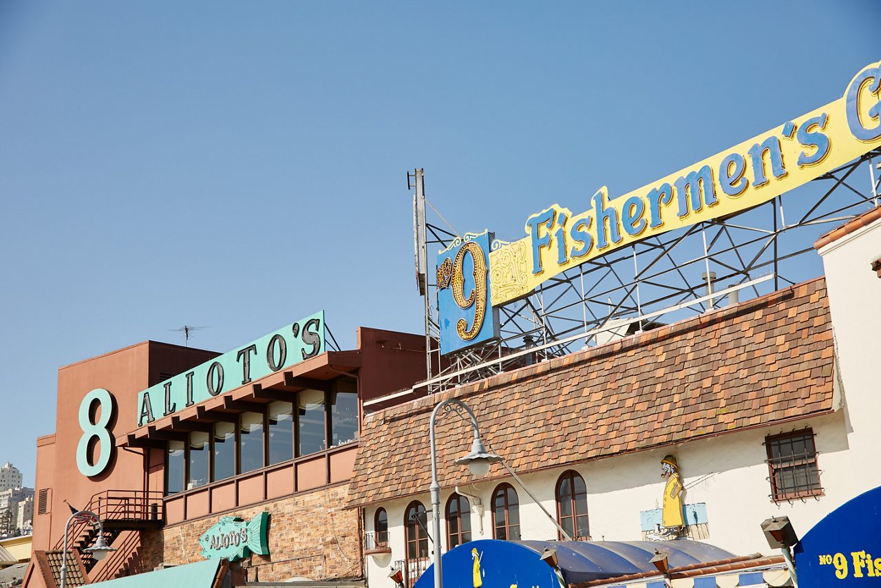 Close-up view of Fisherman's Wharf in San Francisco, showing the Alioto's restaurant sign and the bright yellow Fishermen's Grotto sign against a clear sky..