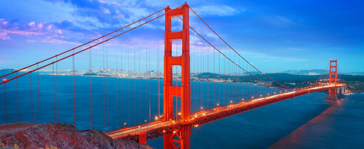 Golden Gate Bridge at dusk with lights on, stretching across the San Francisco Bay, and the city skyline in the background under a vibrant sky.