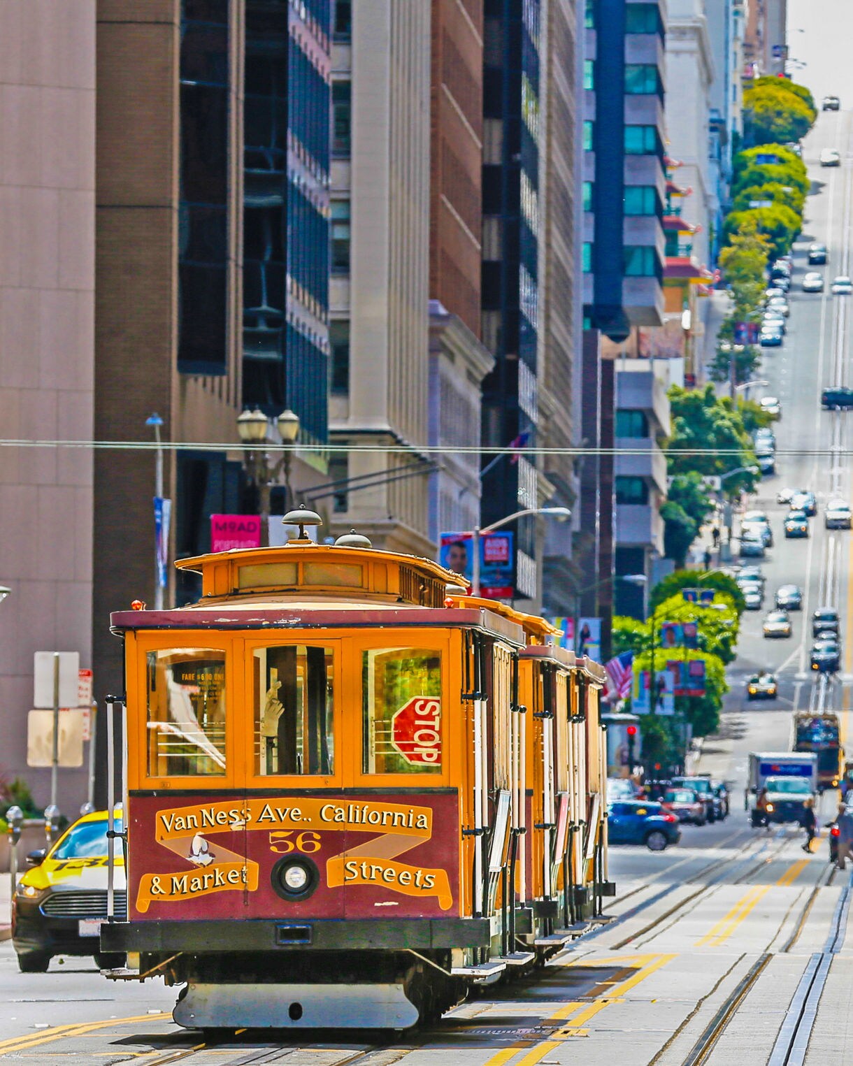 A classic San Francisco cable car numbered 56 travels down a steep hill on Van Ness Avenue, with tall buildings lining the street. The bustling urban scene features cars and pedestrians in the background, capturing the iconic city vibe.