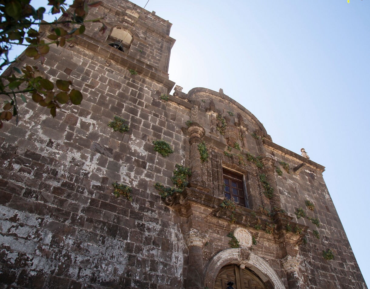 Low-angle view of San Javier Mission in Mexico, showing weathered stone walls, arched details and patches of greenery growing on the façade.