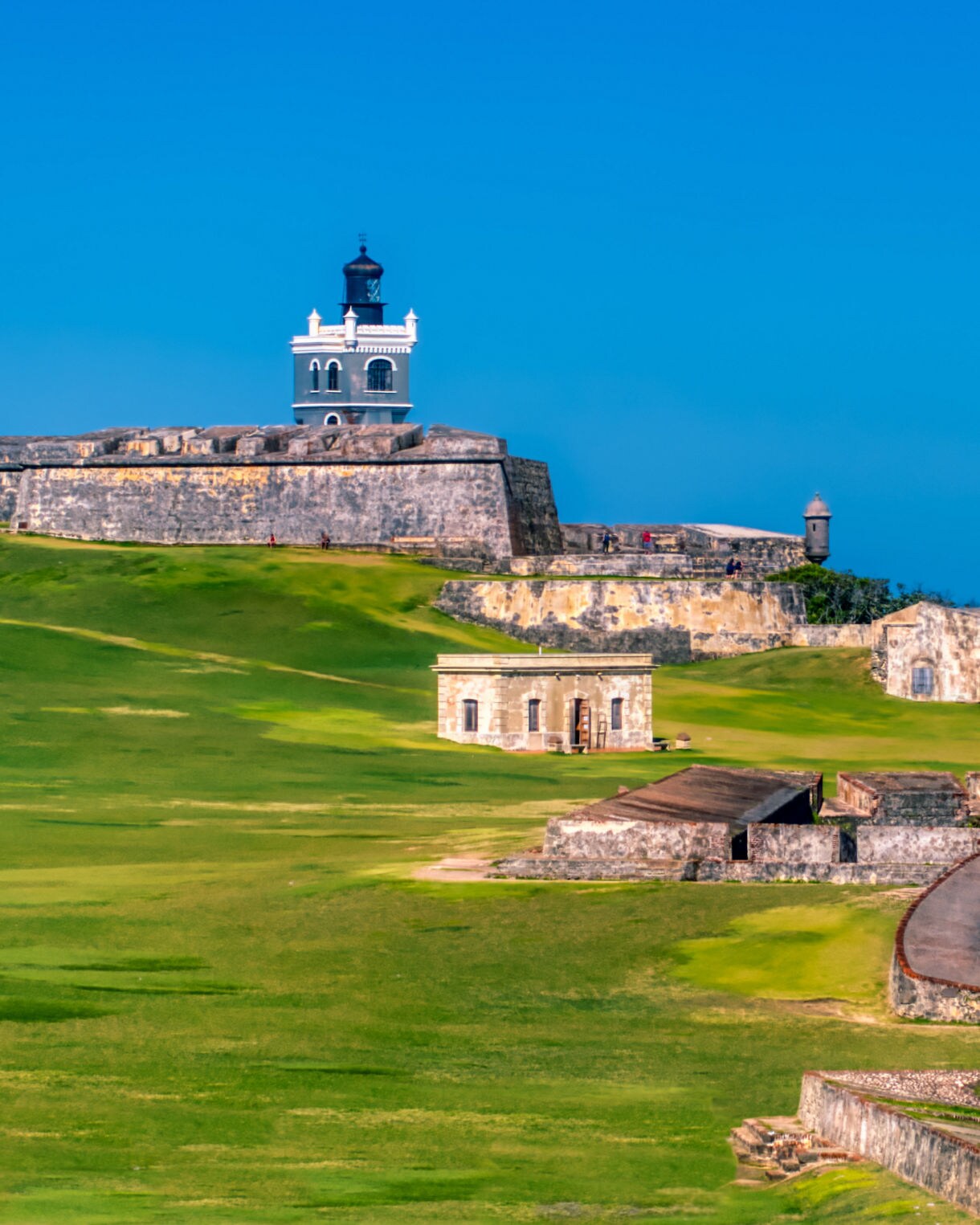 Wide view of Castillo San Felipe del Morro in Old San Juan, showing the fort’s stone walls, lighthouse, grassy hills and the pink-domed cemetery building beside the blue ocean.