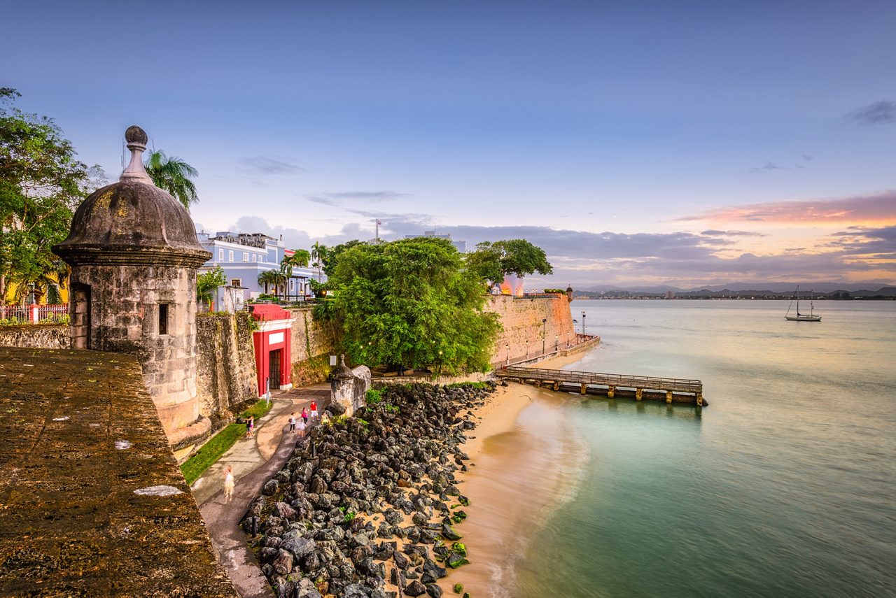 The famous San Juan Gate along Paseo de la Princesa, surrounded by tropical greenery and the sparkling coastline.