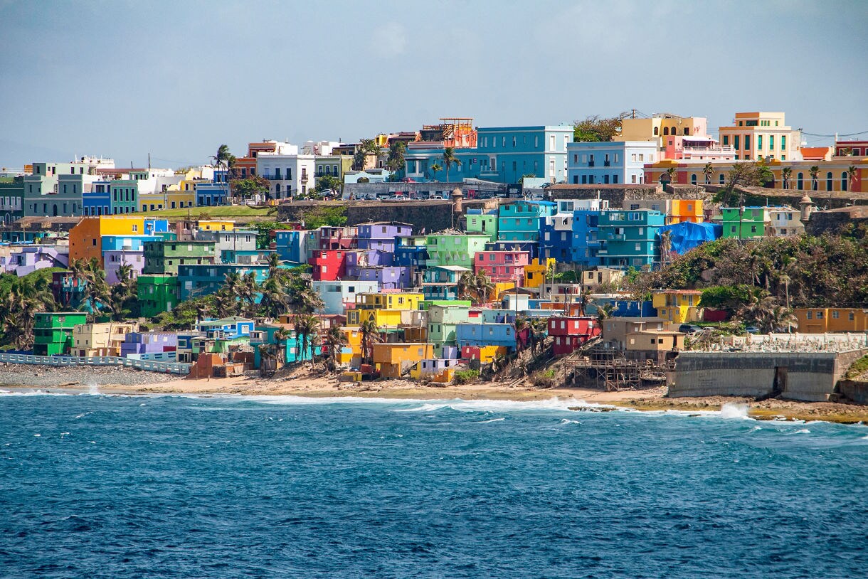 View of San Juan’s vibrant coastal neighborhood with clusters of brightly painted houses in blues, greens, yellows and reds perched on a hillside above the ocean.