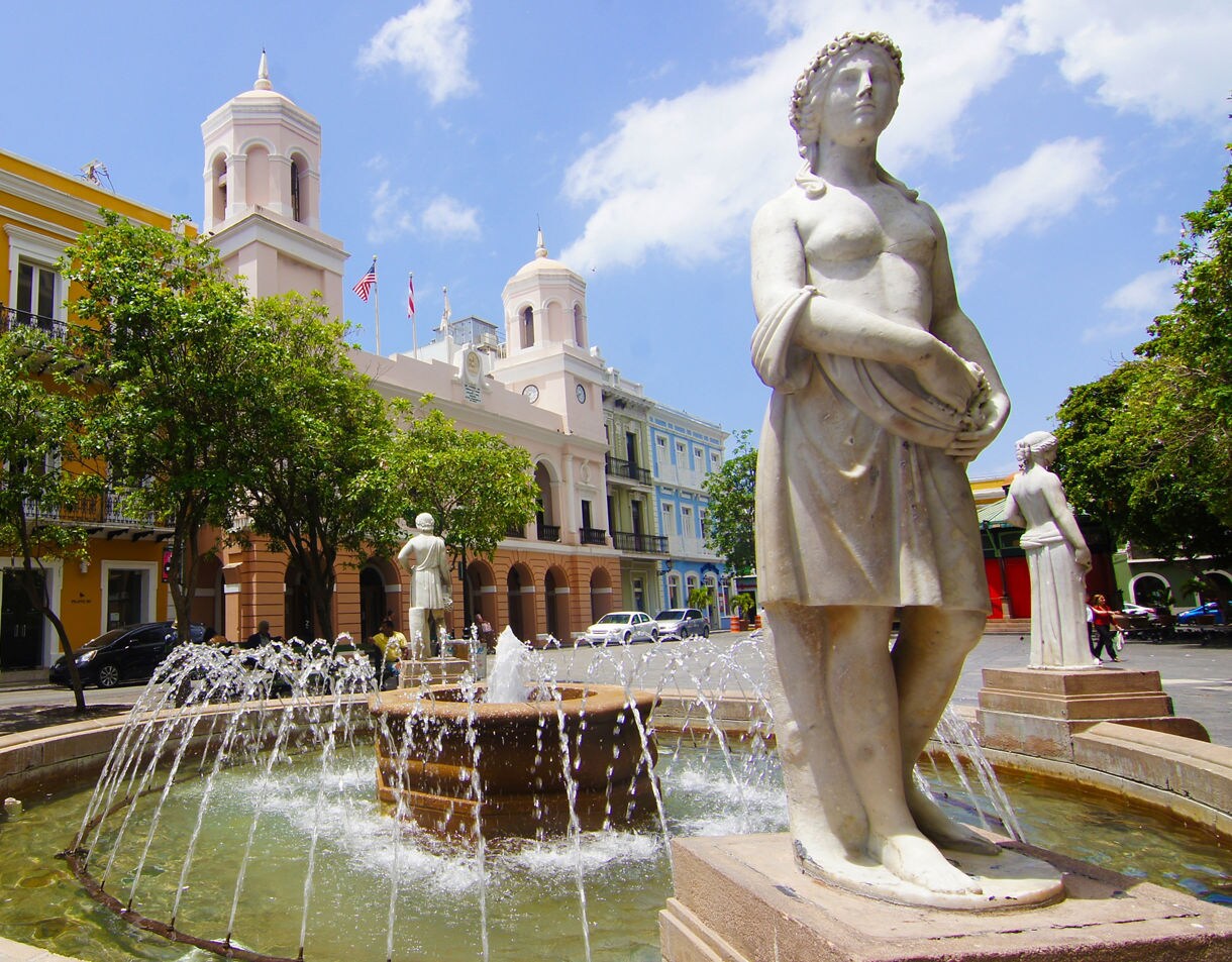 A historic plaza in Old San Juan featuring a circular fountain with classical marble statues. Water sprays from the fountain, framed by colorful colonial buildings in yellow, pink, and blue, along with leafy trees and a bright blue sky.