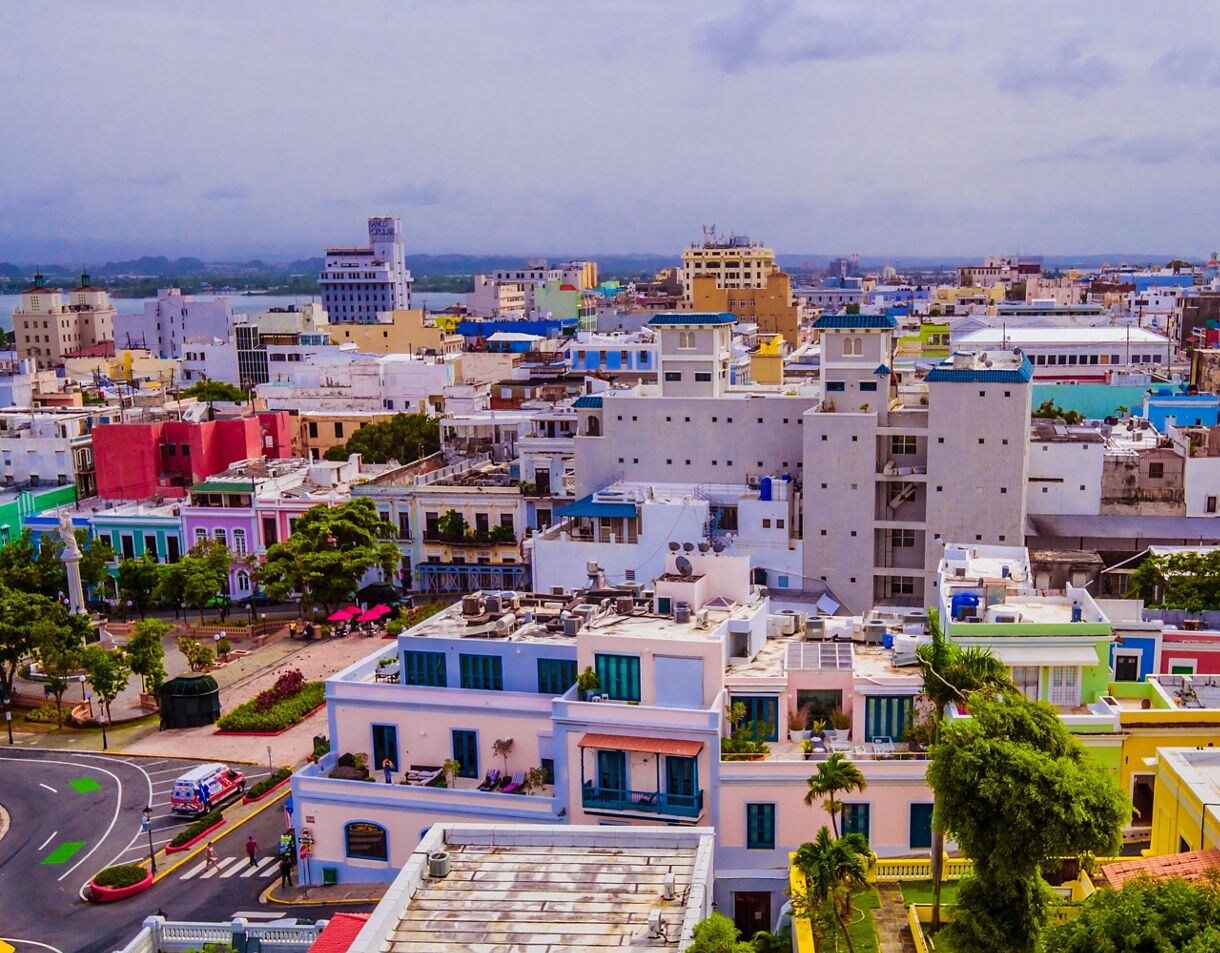 An aerial view of Old San Juan, Puerto Rico, showing a dense cluster of colorful buildings, leafy plazas, and narrow streets. Bright facades in pink, blue, yellow, and green create a vibrant cityscape beneath a cloudy sky.