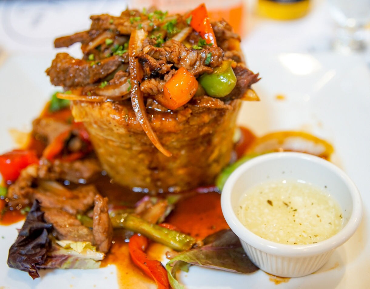 Close-up of a Puerto Rican mofongo dish shaped into a tall mound and topped with shredded beef, peppers and onions. A small cup of garlic sauce sits to the side on a white plate.