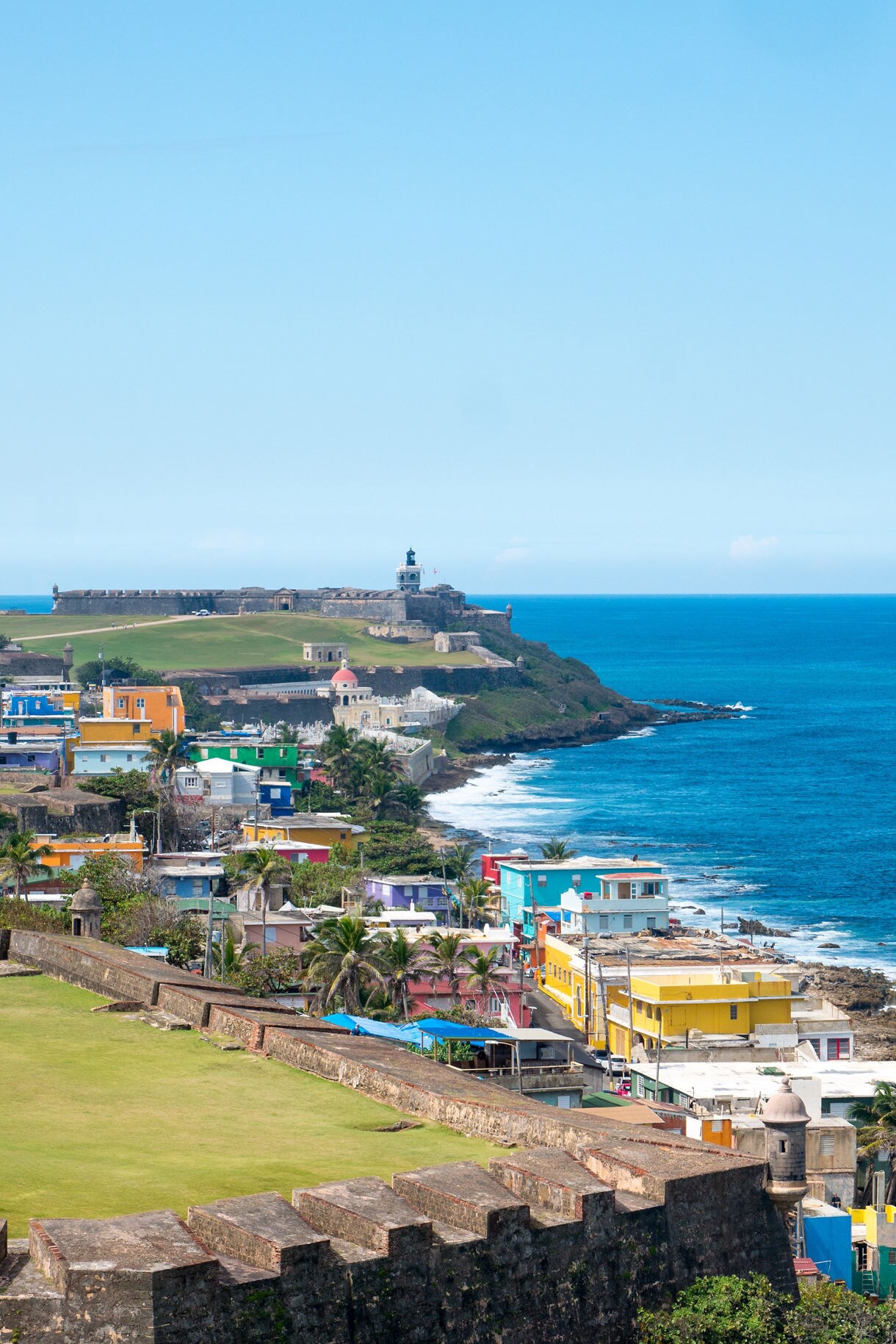 Vertical photo of Old San Juan’s coastline with colorful houses in La Perla in the foreground and the stone walls and lighthouse of Castillo San Felipe del Morro overlooking the bright blue ocean in the distance.