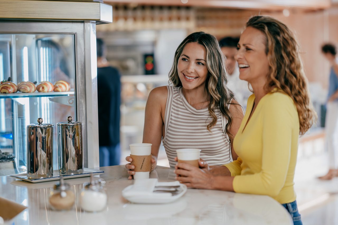 Two women enjoying coffee together at a coffee bar counter, with one wearing a striped top and the other in a yellow sweater, pastry display case visible beside them.