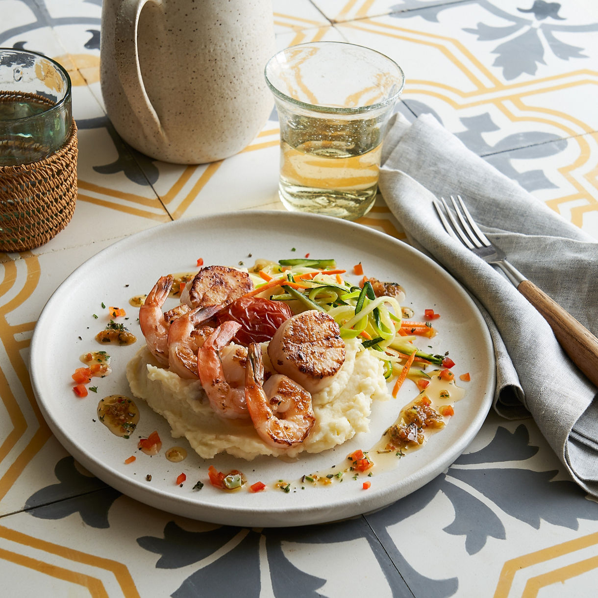 A plated dish of creamy pasta topped with shrimp and garnished with herbs, served on a white plate atop a patterned tablecloth with a glass of wine nearby.