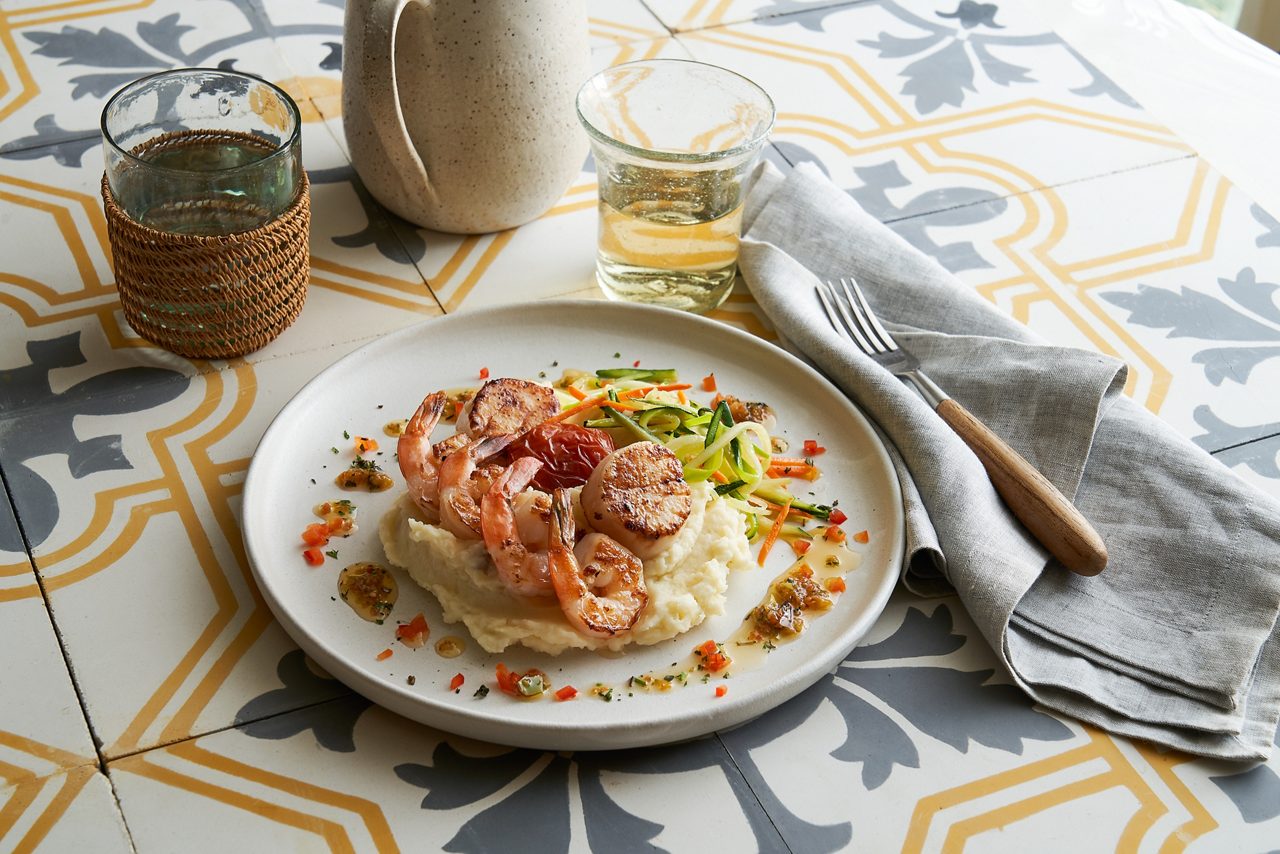 A plated dish of creamy pasta topped with shrimp and garnished with herbs, served on a white plate atop a patterned tablecloth with a glass of wine nearby.