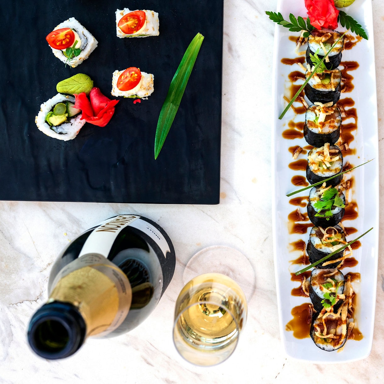 A stylized overhead view of three sushi-type dishes arranged against dark plates - assorted nigiri pieces, a long dragon roll, and colorful appetizers, with a bottle and glass of wine in the bottom corner. 