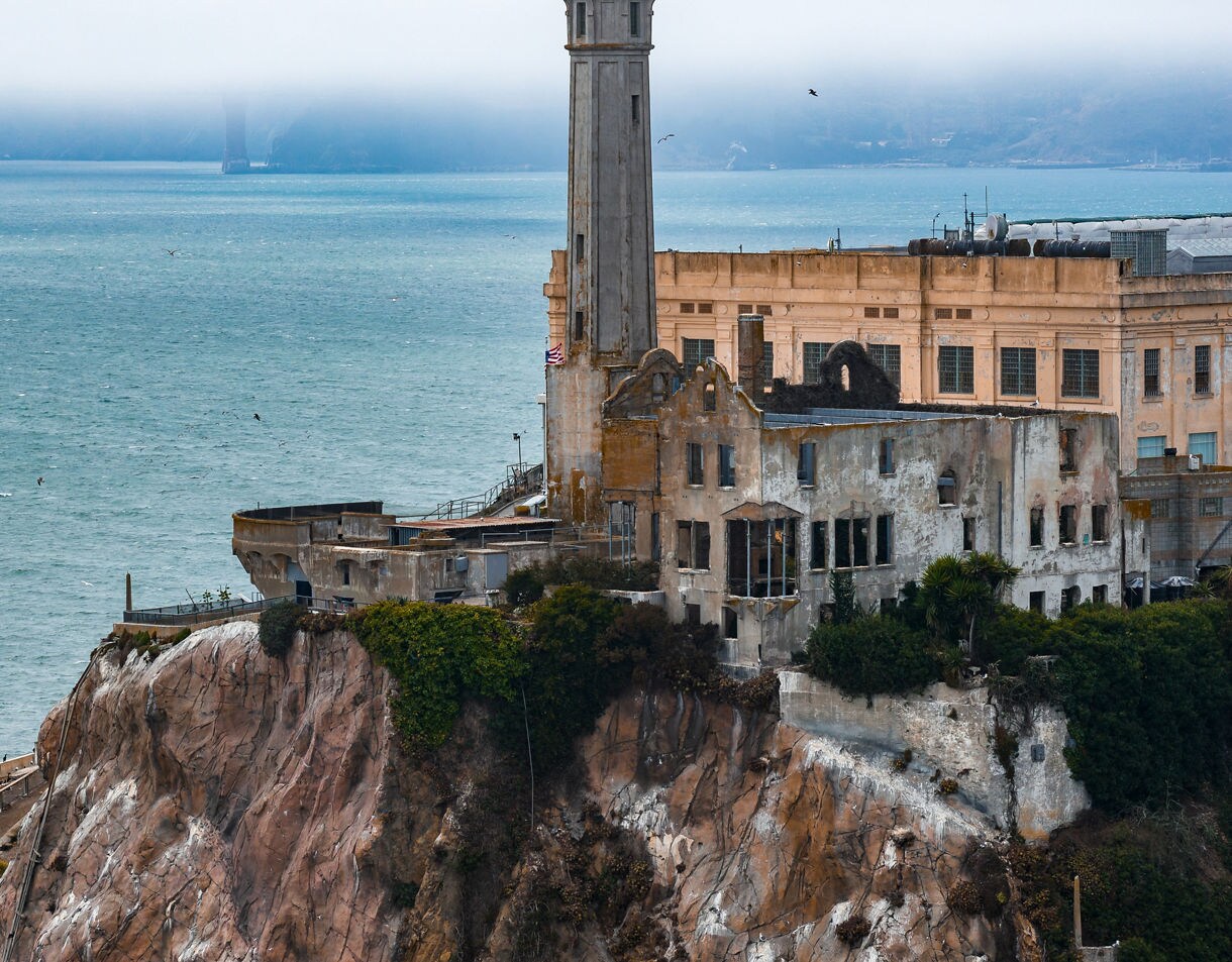 A close view of Alcatraz Island’s weathered prison buildings perched on rocky cliffs with peeling walls, overgrown plants and the surrounding blue water under a layer of low fog.