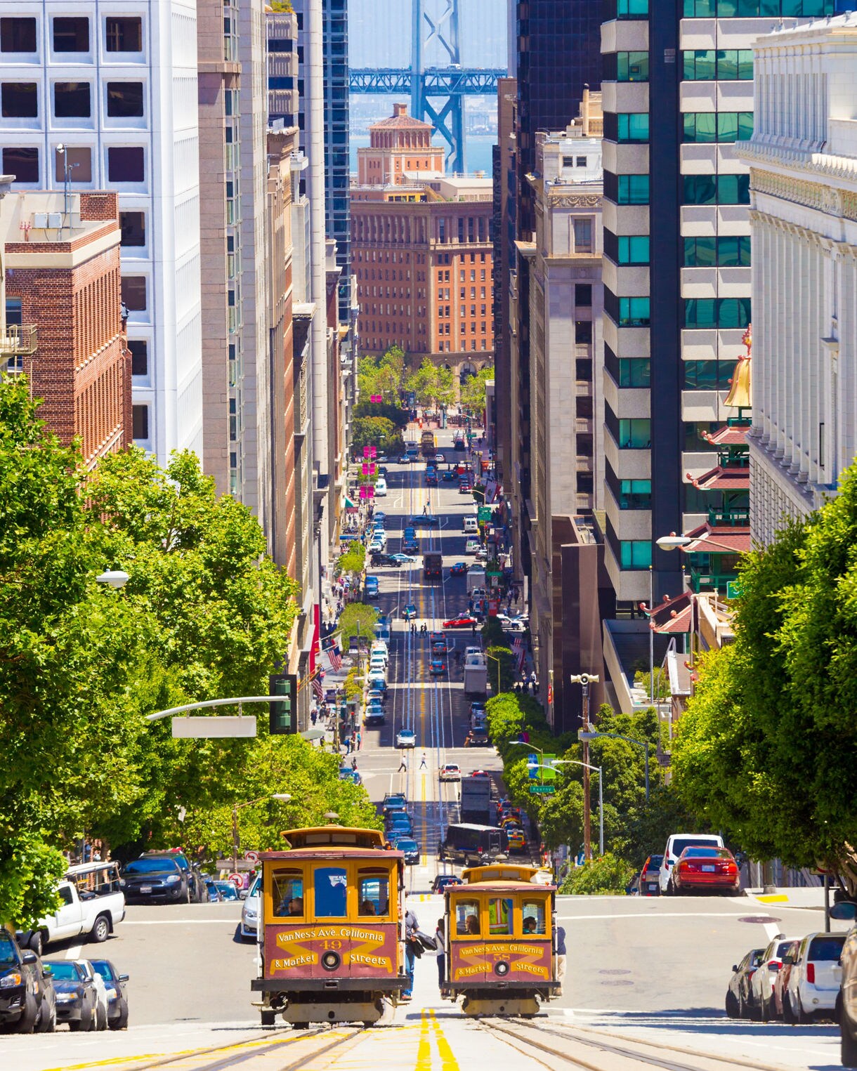 A steep San Francisco street with two cable cars ascending the hill, tall buildings lining both sides, busy traffic below and the Bay Bridge visible in the background through the corridor of skyscrapers.