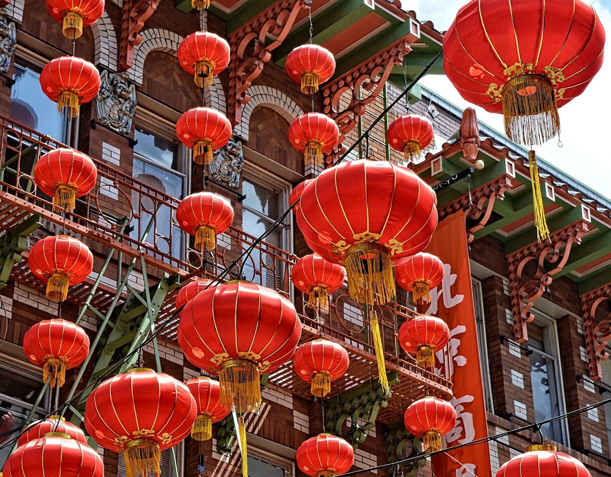 A close-up view of bright red Chinese lanterns hanging above a Chinatown street with ornate building facades, decorative trim and fire escapes in the background.