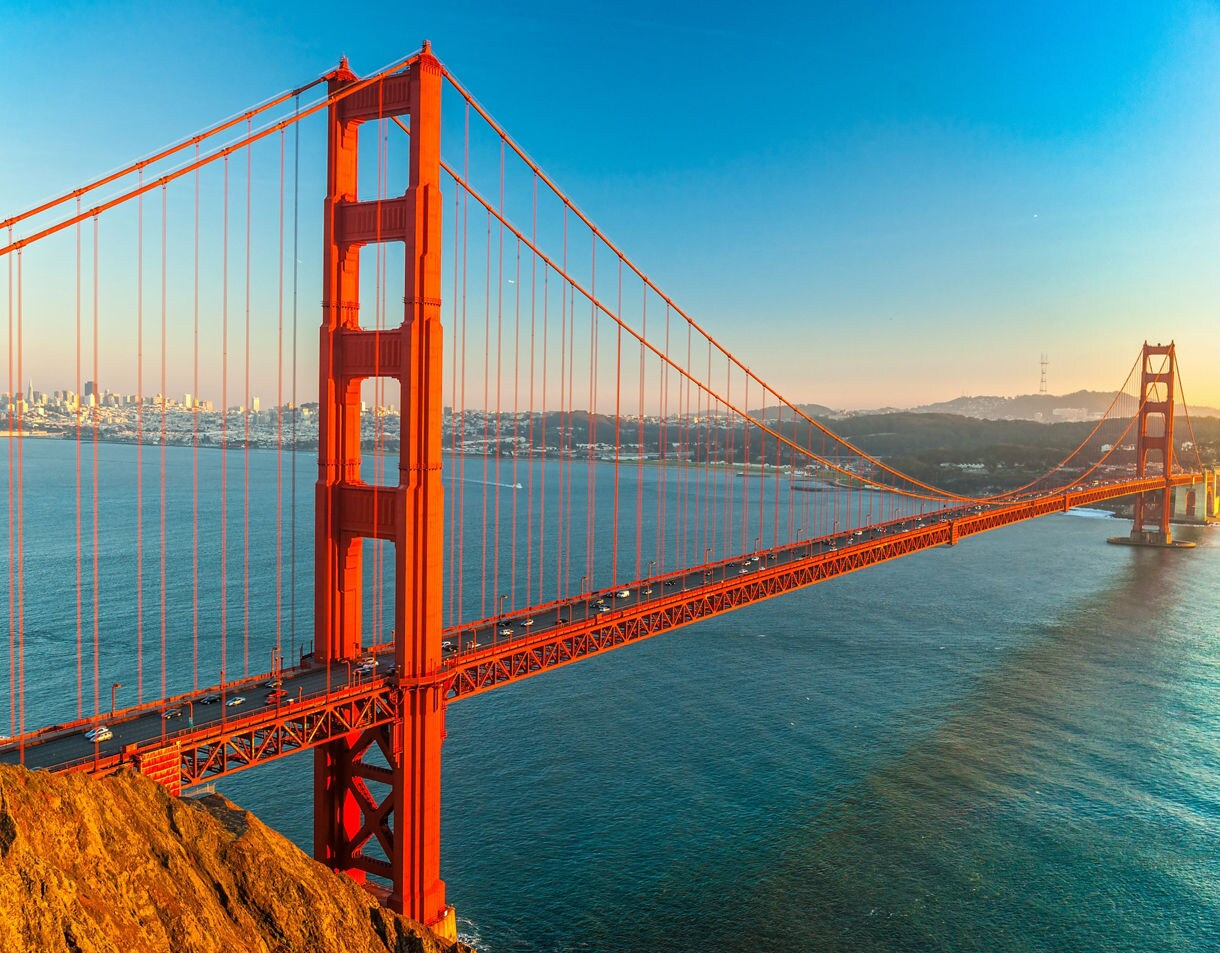 A wide-angle view of the Golden Gate Bridge at sunset showing its bright orange towers and cables stretching over the blue waters of the San Francisco Bay with the city skyline in the distance.