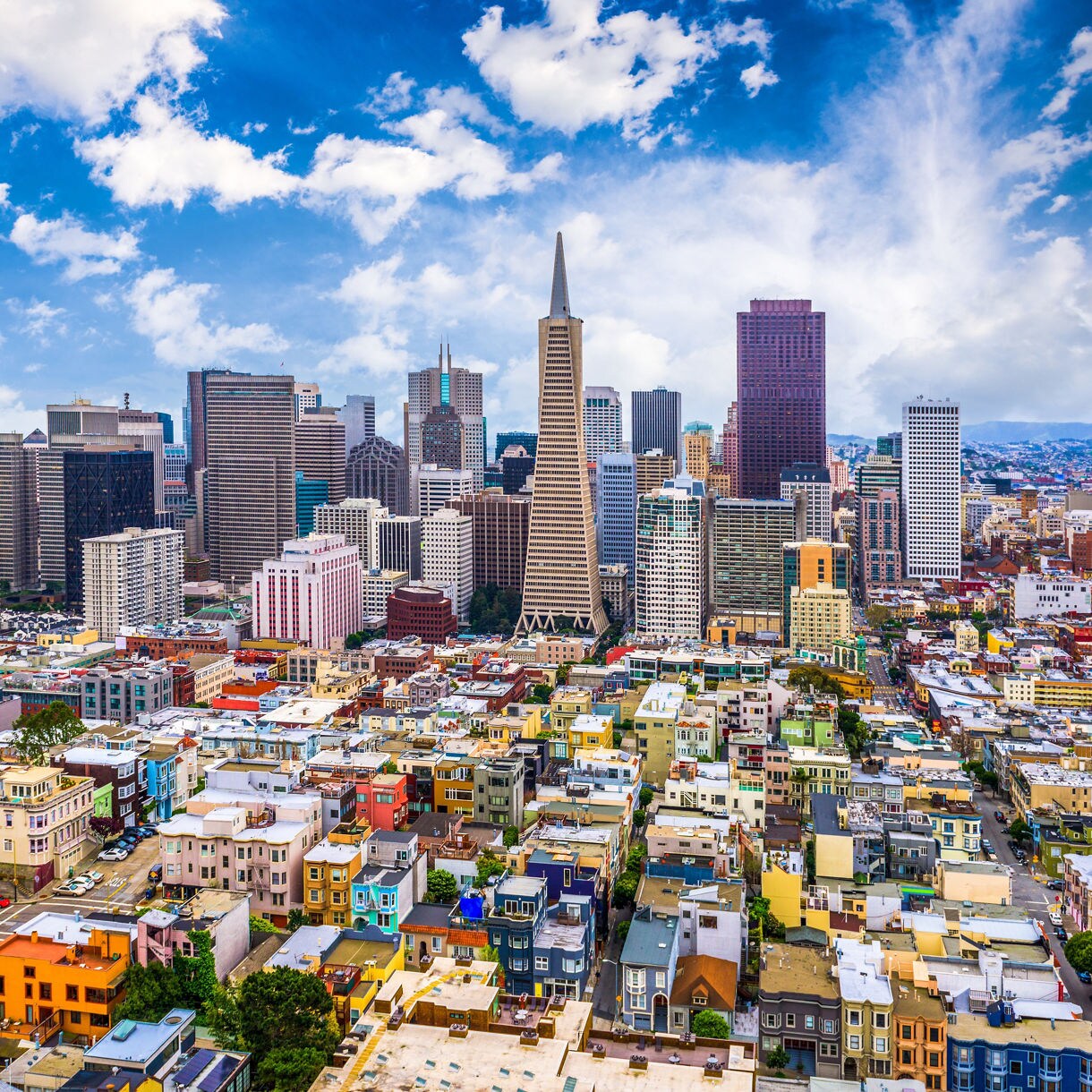 A vibrant aerial view of San Francisco showing rows of colorful homes in the foreground and the downtown skyline with the Transamerica Pyramid, high-rise buildings and the Bay Bridge under a bright blue sky with scattered clouds.