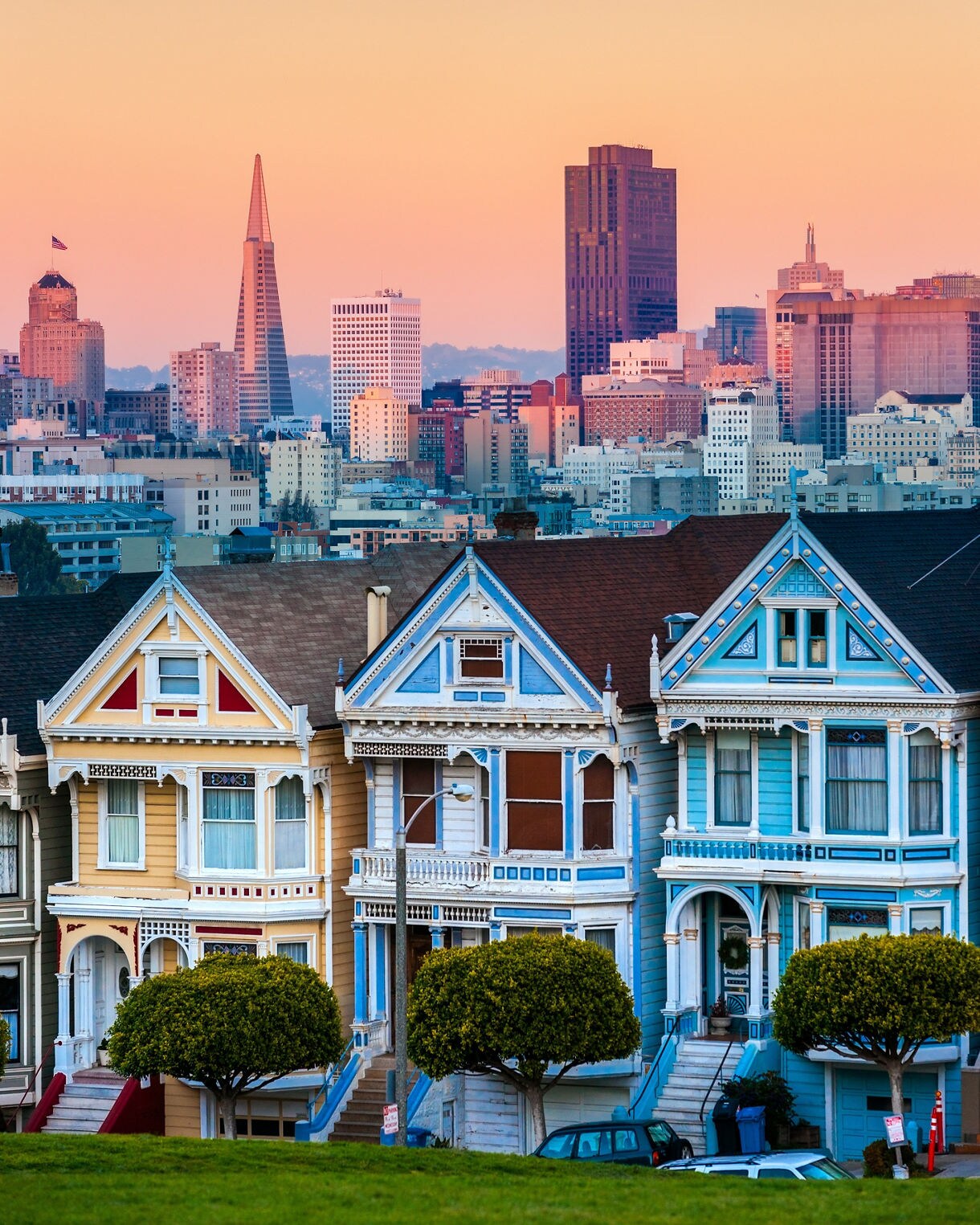 A view of San Francisco’s Painted Ladies, a row of colorful Victorian houses in the foreground with the city skyline behind them including the Transamerica Pyramid and tall buildings under a warm sunset sky.