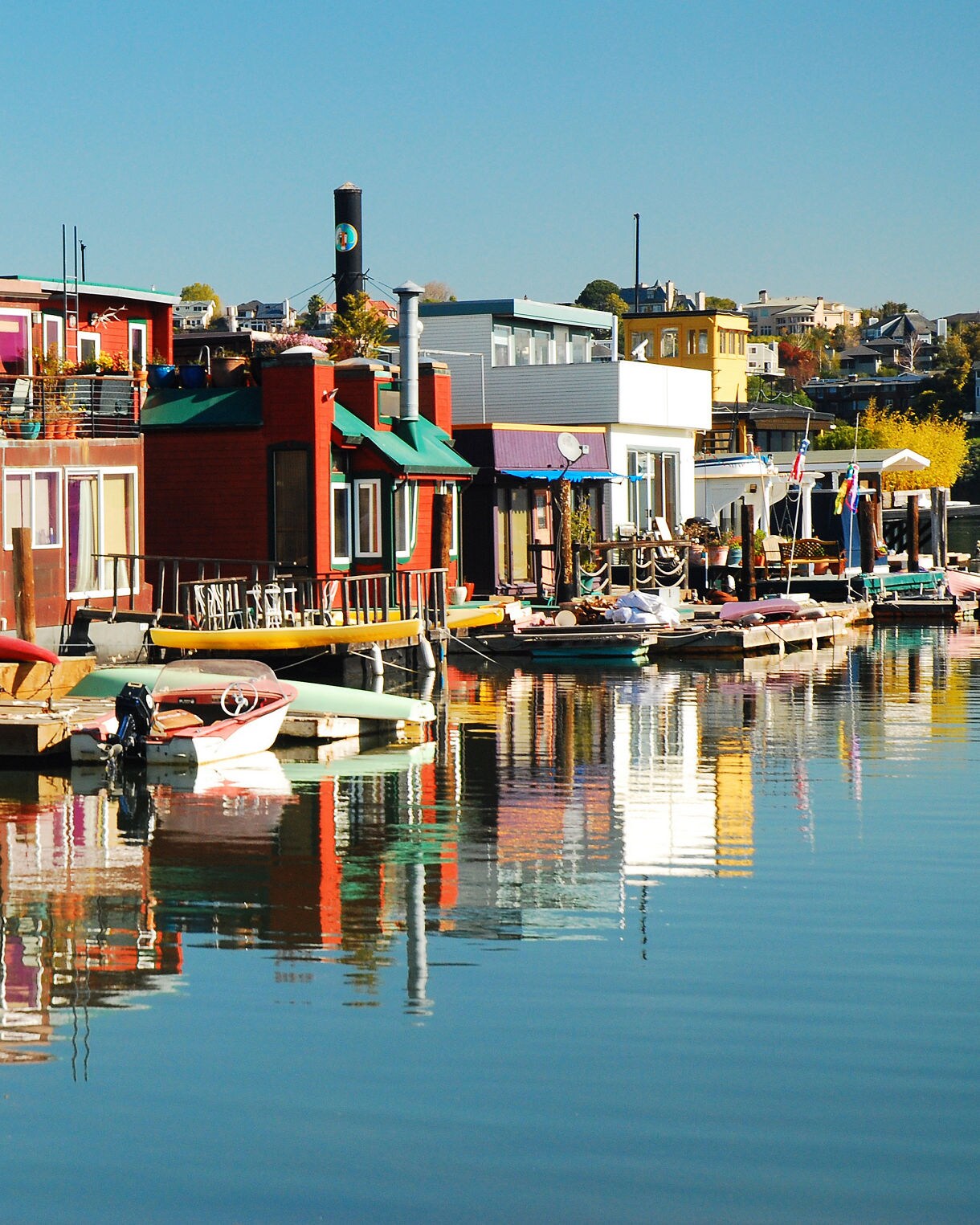 A line of colorful Sausalito houseboats sitting along a calm marina with wooden decks, small boats moored nearby and vivid reflections of the homes shimmering on the water under a clear blue sky.