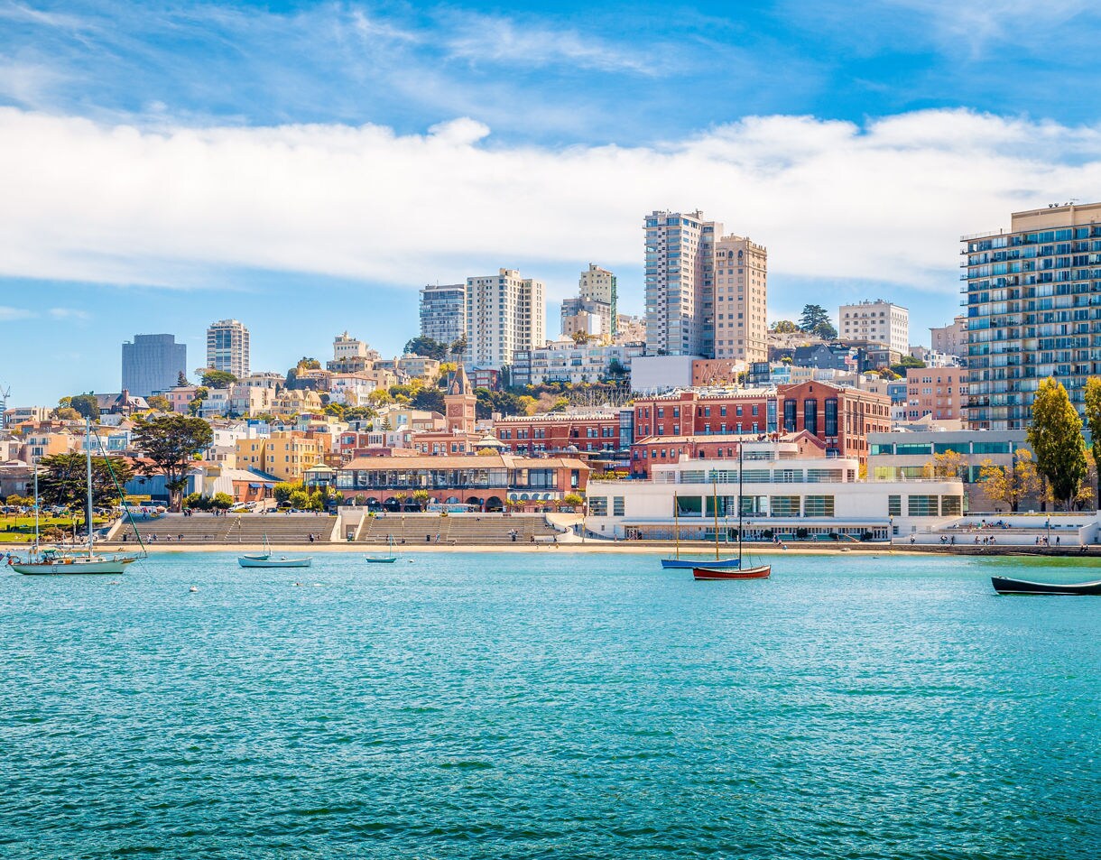 A scenic view of San Francisco’s waterfront with small boats floating on bright blue water and a backdrop of colorful buildings, brick structures and high-rise towers climbing the hillside under a clear sky.