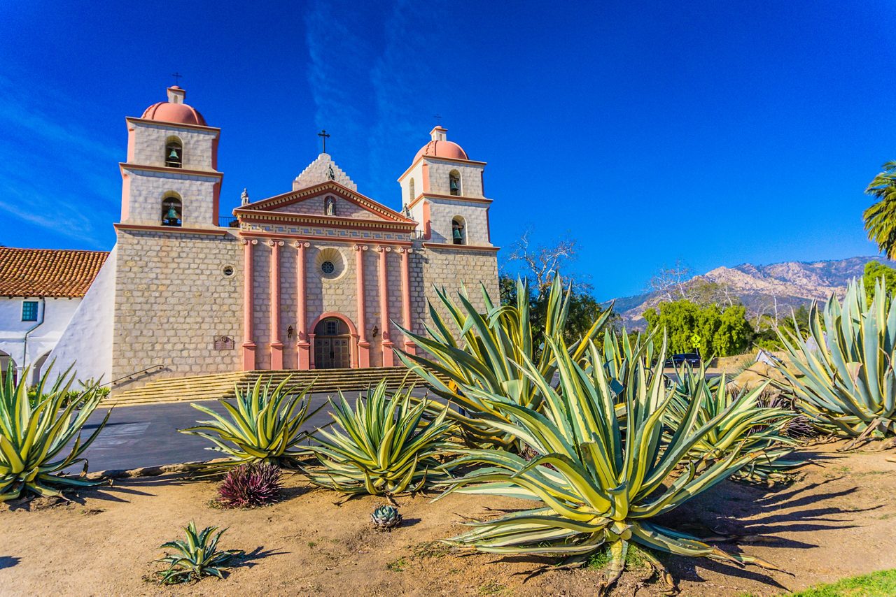 The Old Mission in Santa Barbara, California, featuring its stone facade and twin bell towers, surrounded by large agave plants, under a clear blue sky with mountains in the background.