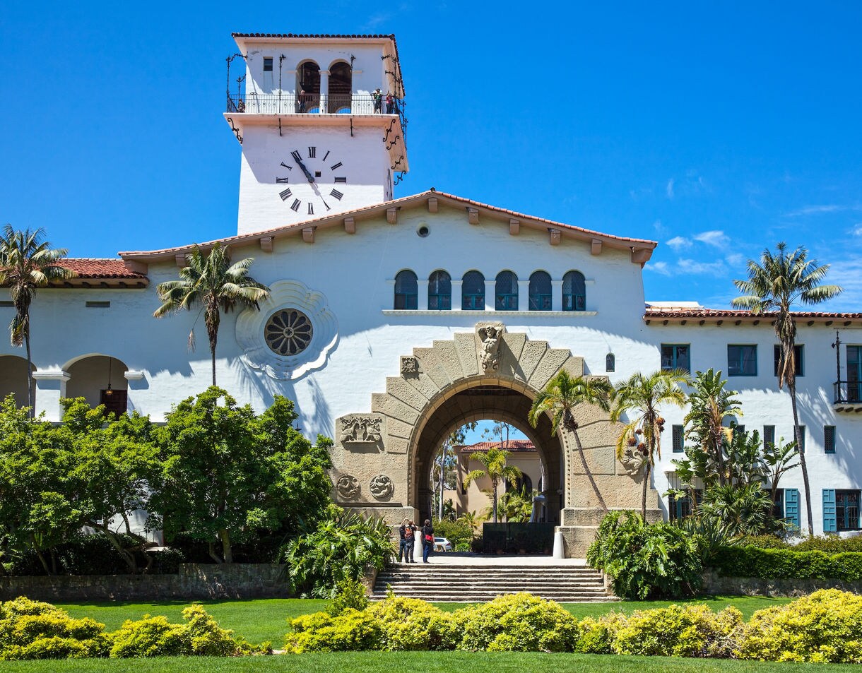 Mission building with weathered arched colonnade, bell tower, and palm trees along a paved walkway.